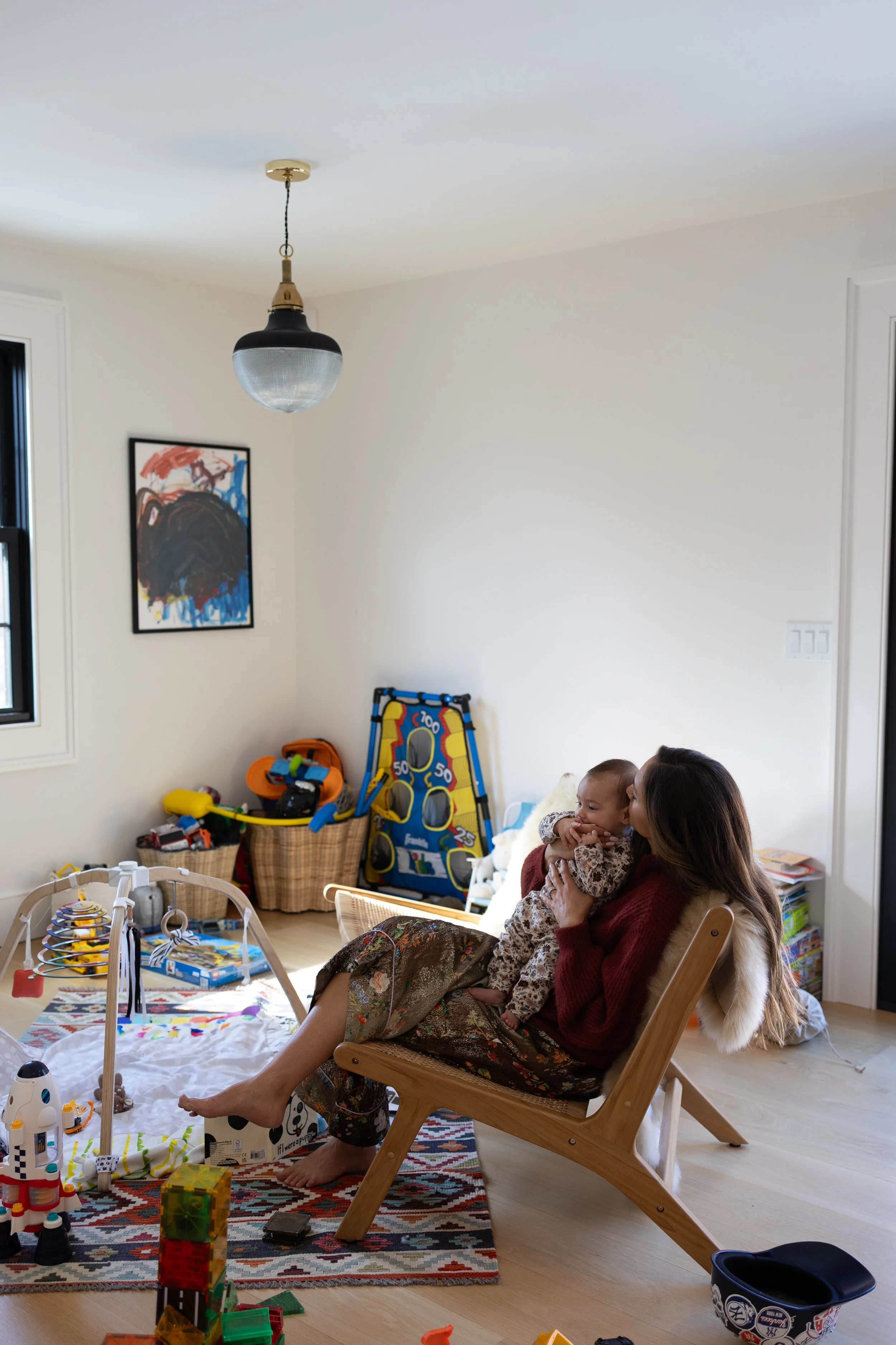 A woman sitting in a wooden chair, holding a baby, in a playroom with toys and game equipment on the floor and a basket of toys in the background.