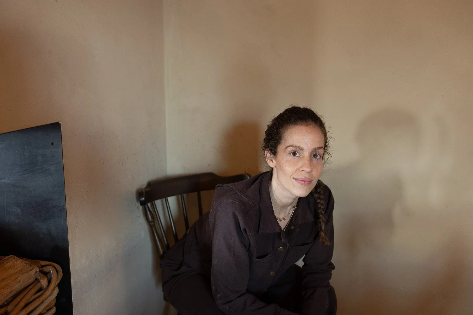 A woman with dark curly hair braided over her shoulder, sitting on a wooden chair, wearing a dark jacket, in a room with beige walls.