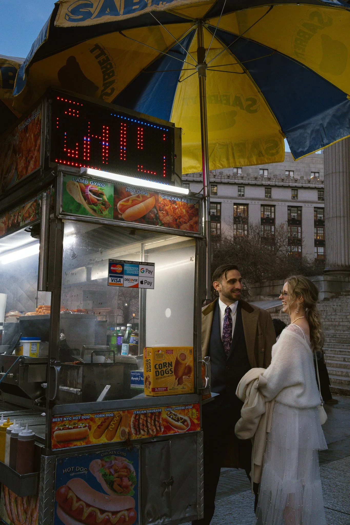 A street food stand selling hot dogs, chicken gyros, and other quick foods, with two people in front of it, a man and a woman, smiling and talking. The stand has a yellow and blue umbrella and a digital sign above it.