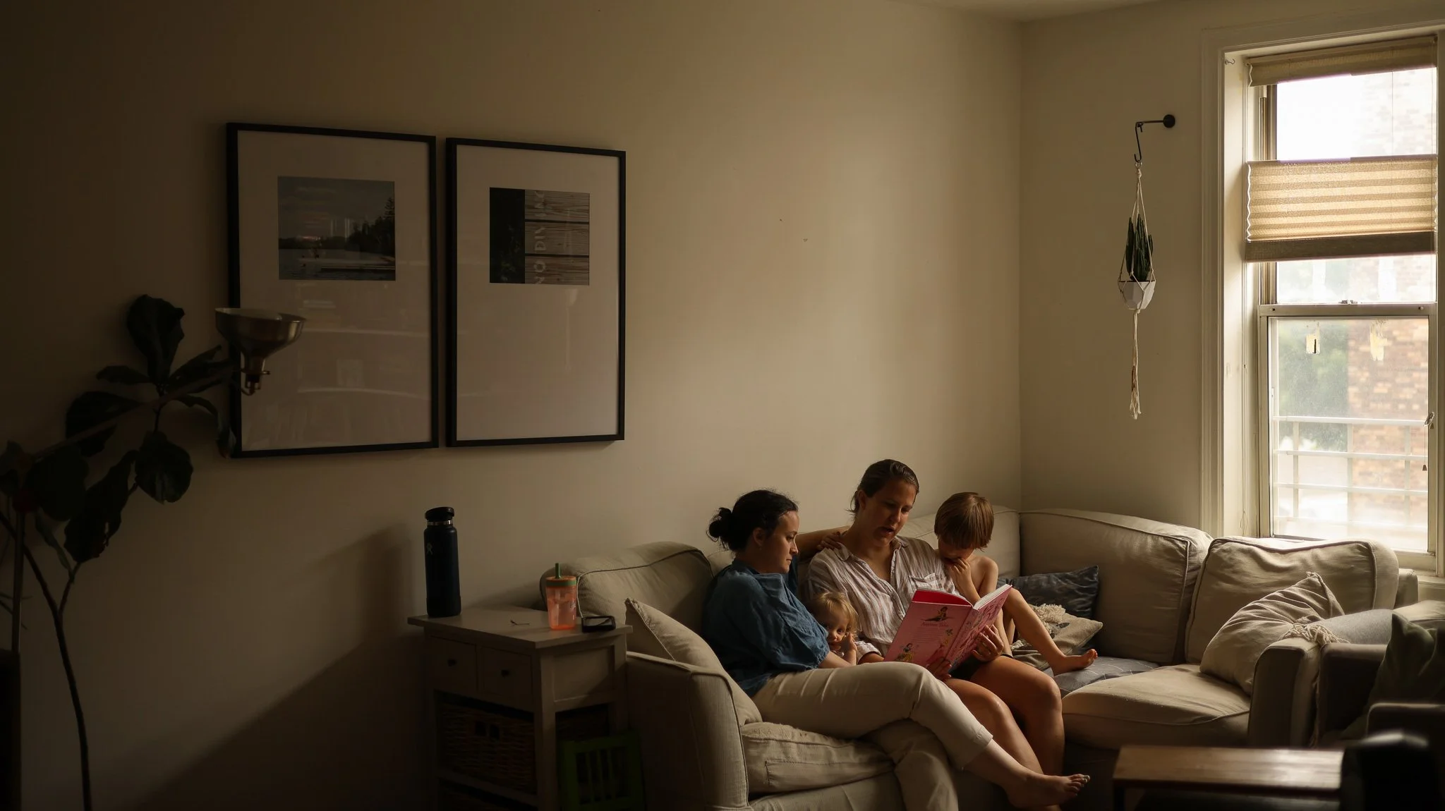 Family sitting on a beige couch in a living room, reading a book together, near a large window with sunlight streaming in, with framed pictures on the wall and a side table with a water bottle.