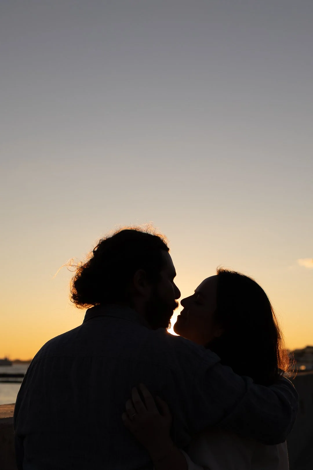 Silhouetted couple embracing at sunset by the water, with a clear sky and a few distant boats or structures.