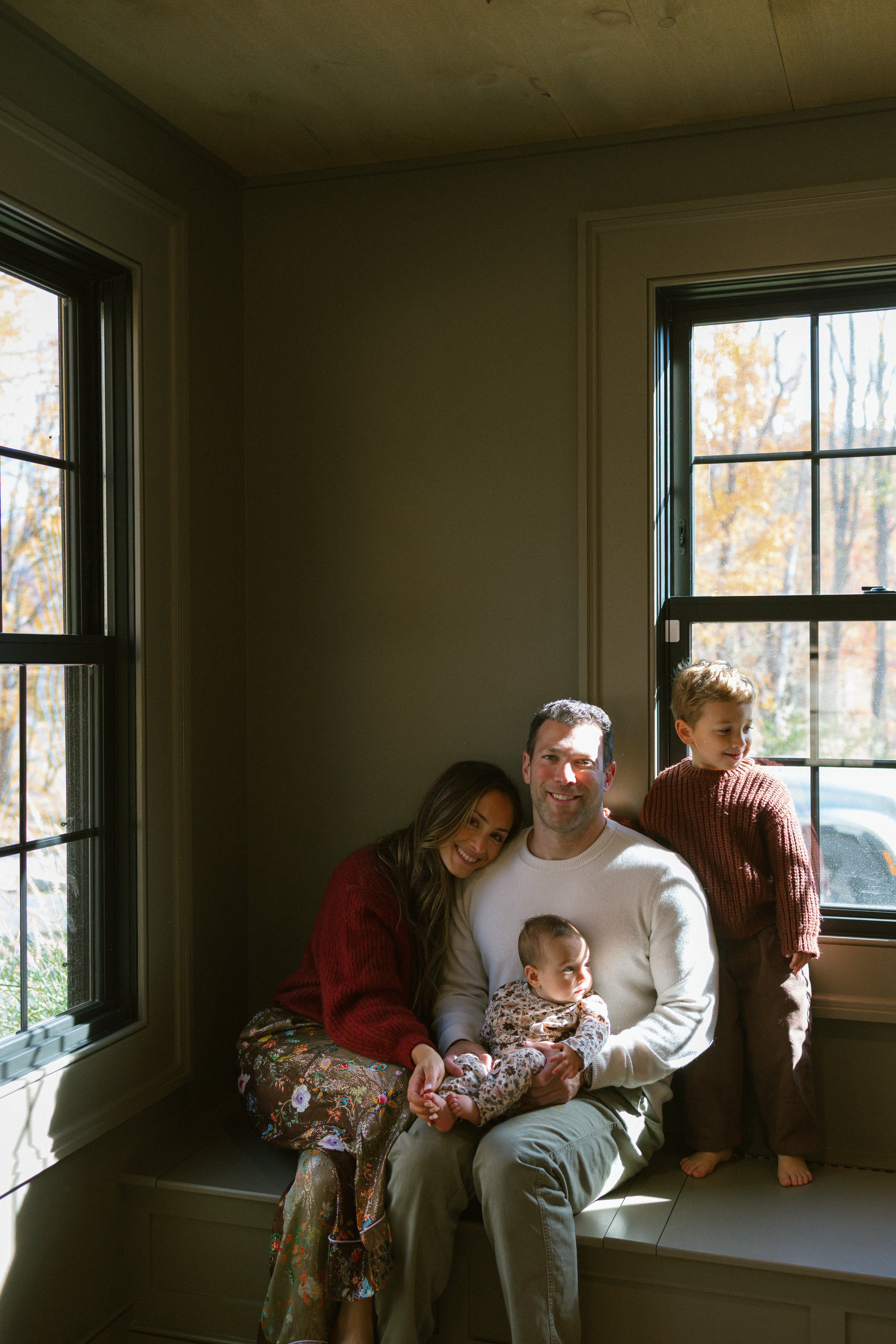 A family of four sitting on a window seat in a sunlit room during fall. Mom, dad, young boy, and baby girl smiling.