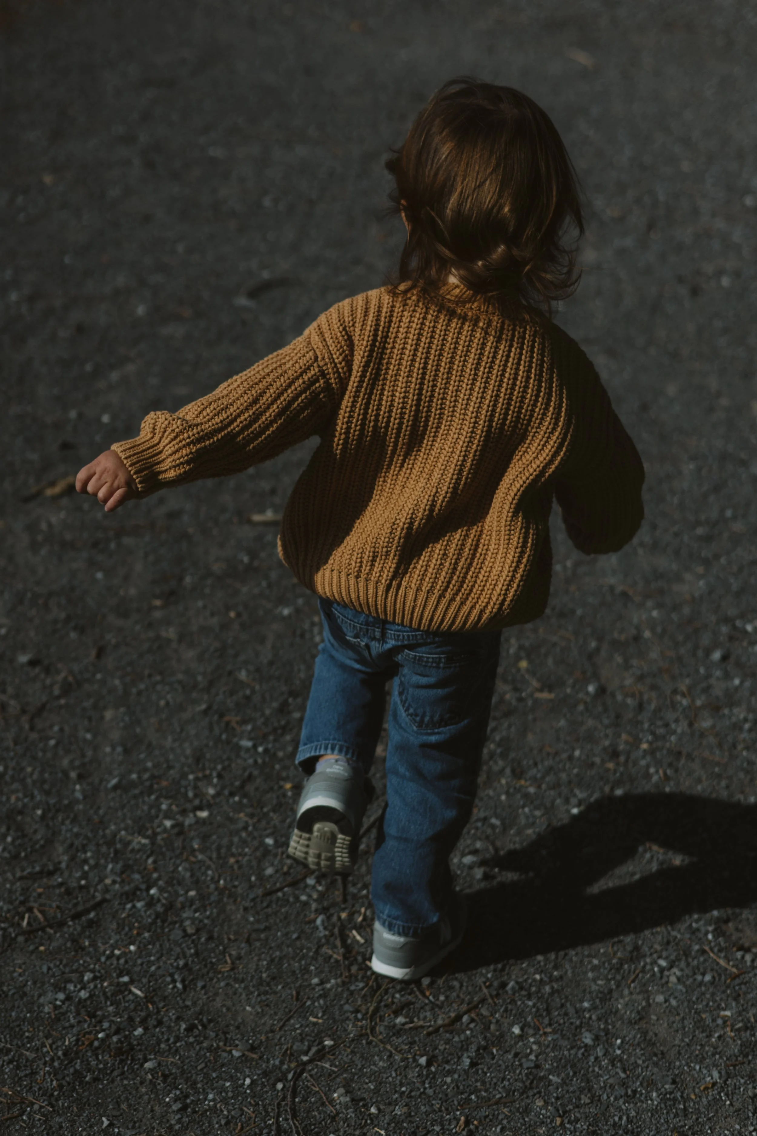 Child in brown sweater and blue jeans walking on dark gravel surface with shadow cast behind.