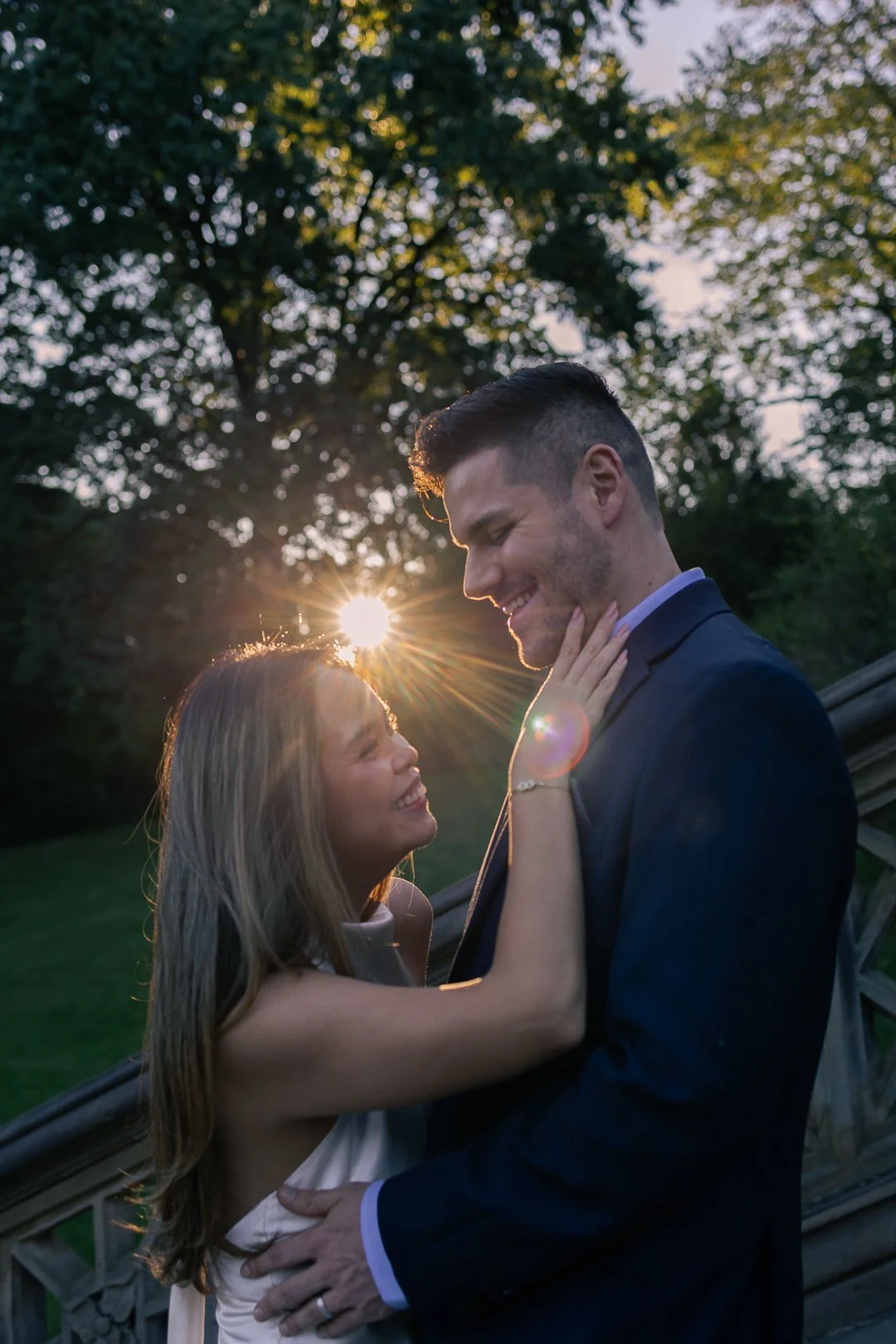 A couple dressed in formal attire smiling and looking at each other outdoors during sunset, with trees in the background.