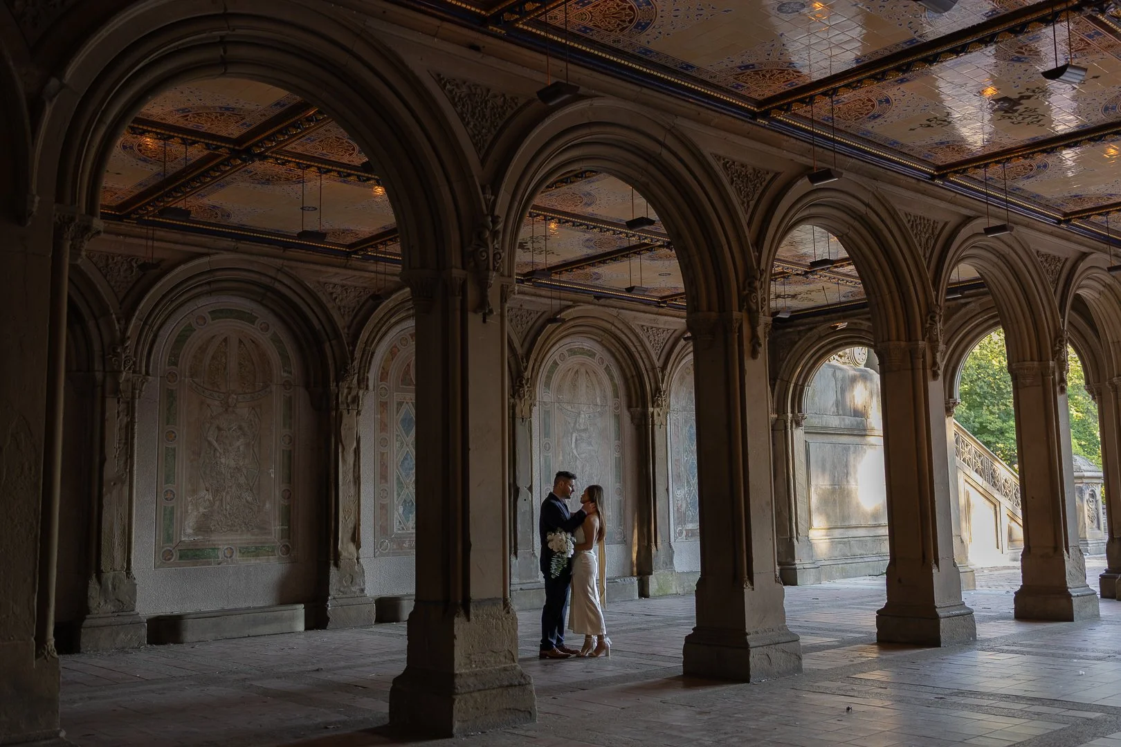 A couple stands under arches in a historic building, with intricate ceiling designs and sunlight streaming through. The man is holding a bouquet.