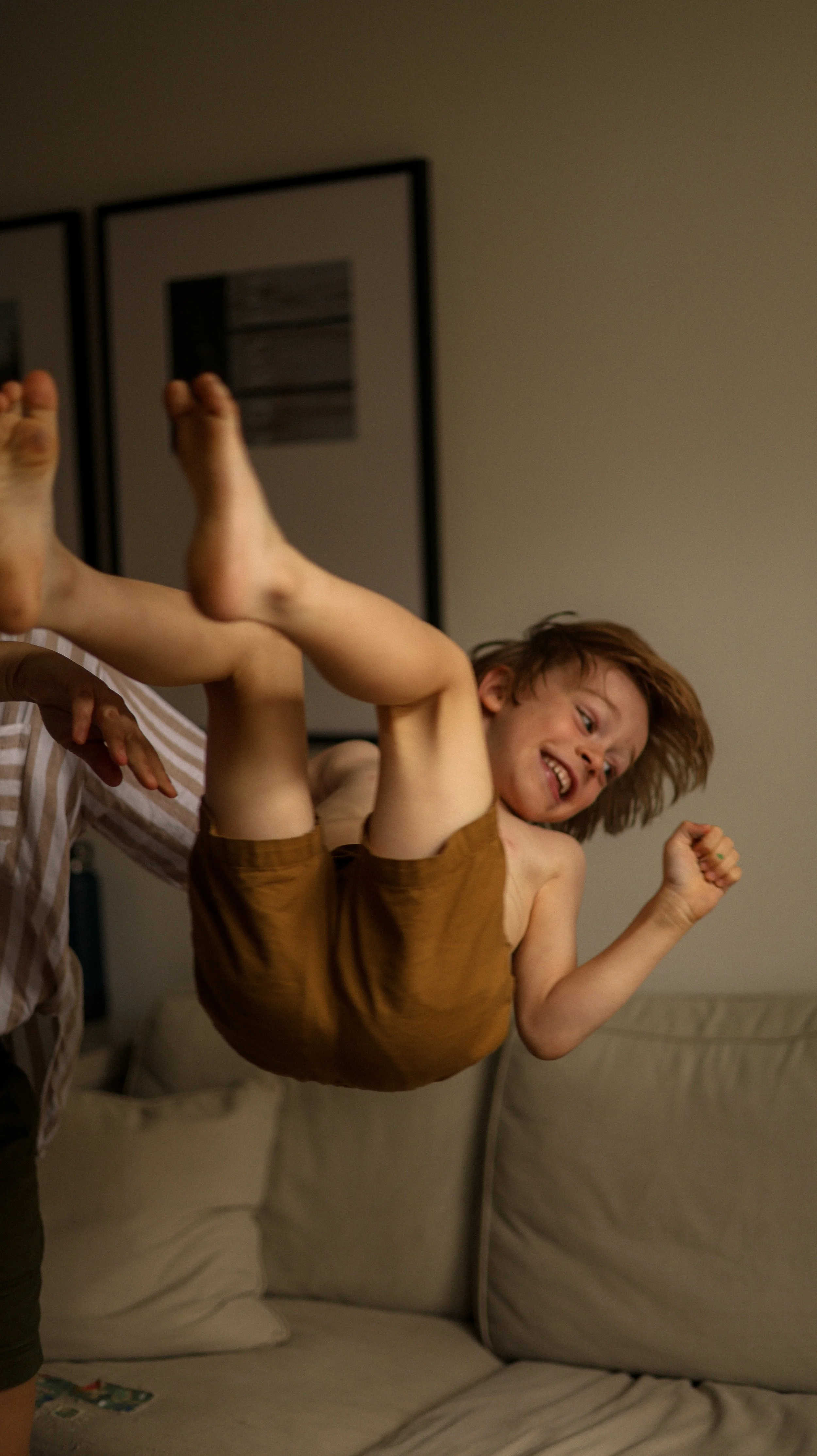 A young boy is swinging in midair, held by an adult, indoors, with a smiling face and tousled brown hair.