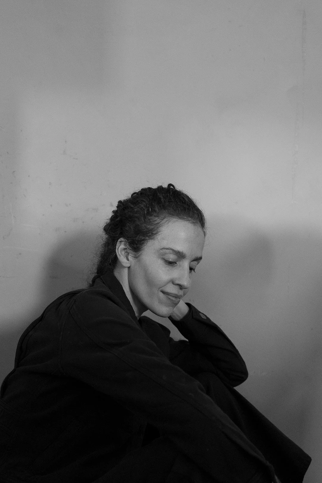 A black and white photo of a woman sitting against a plain wall, with her eyes closed and a serene expression, wearing a dark jacket.