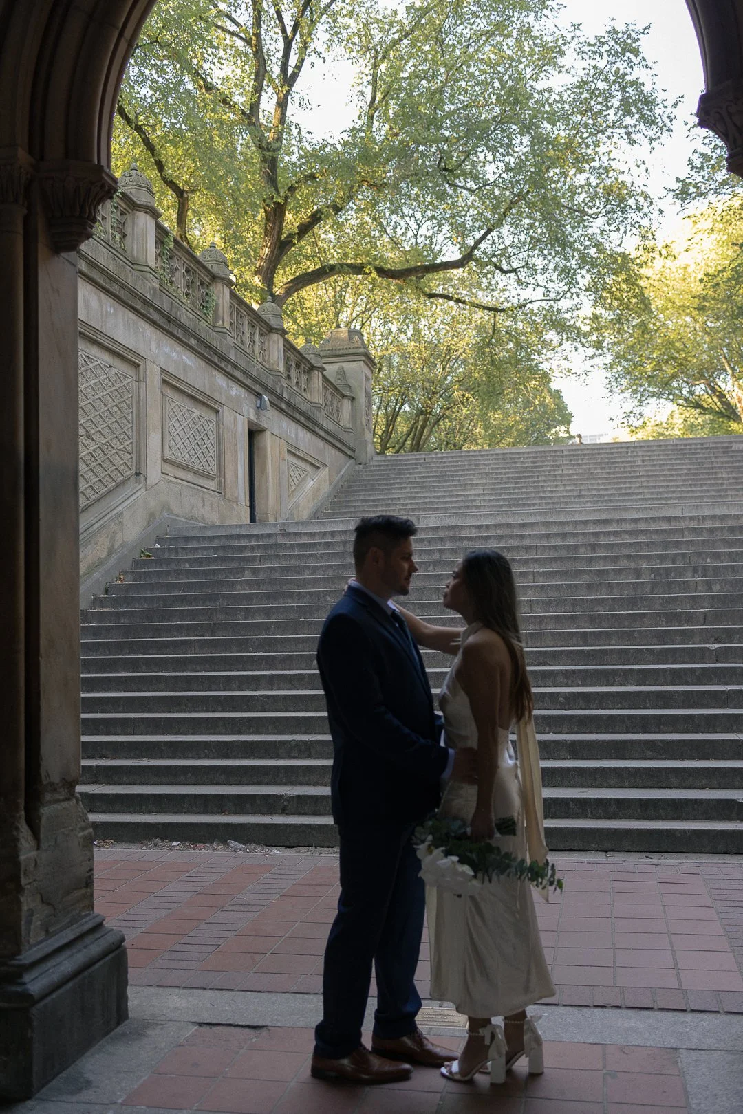 A couple in wedding attire standing under an archway, facing each other, with stairs and trees in the background.