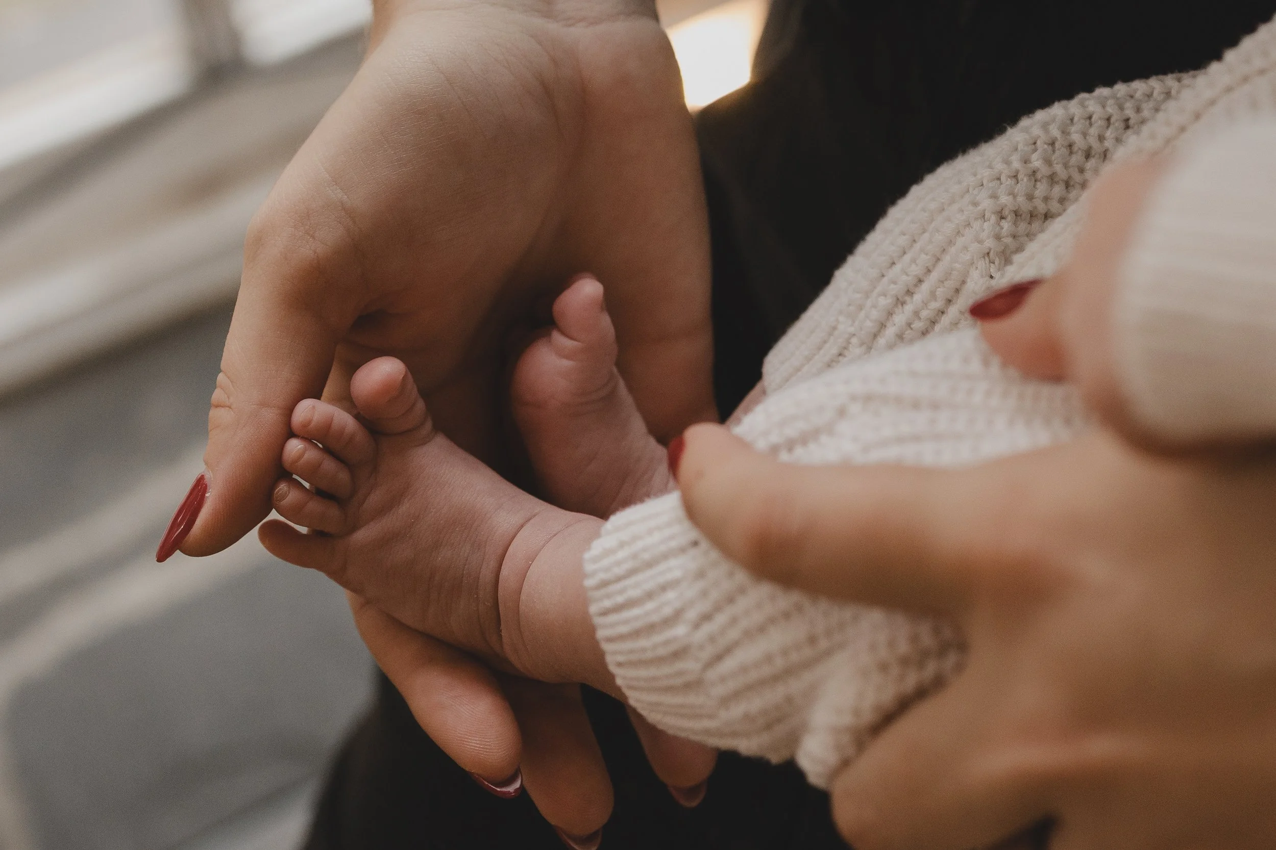 A close-up of a person's hand holding a baby's hand, with the baby's hand resting on its chest, wearing a beige knitted sweater.