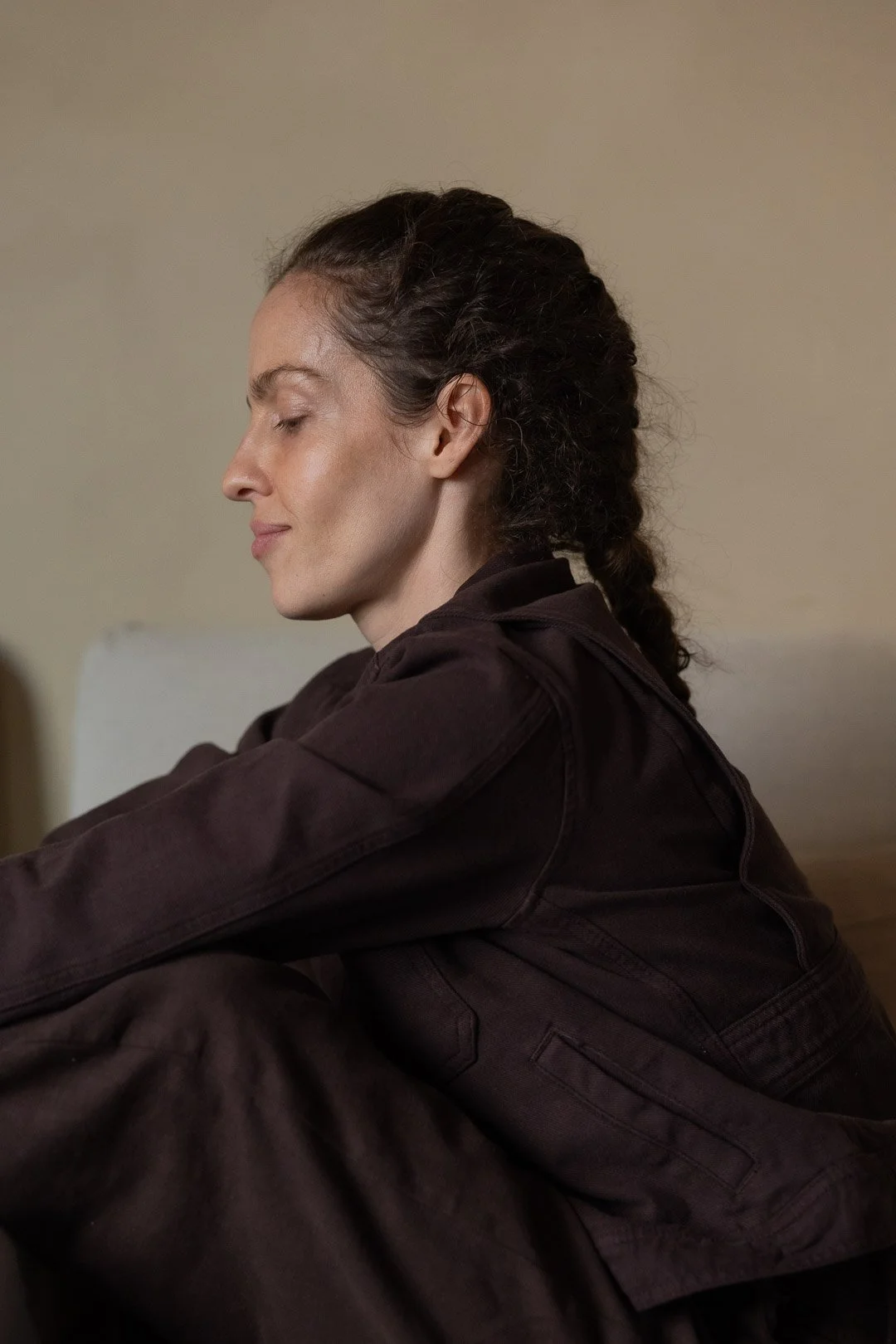 Profile of a woman with dark, curly hair in a braid, sitting with eyes closed, wearing a dark jacket against a neutral background.