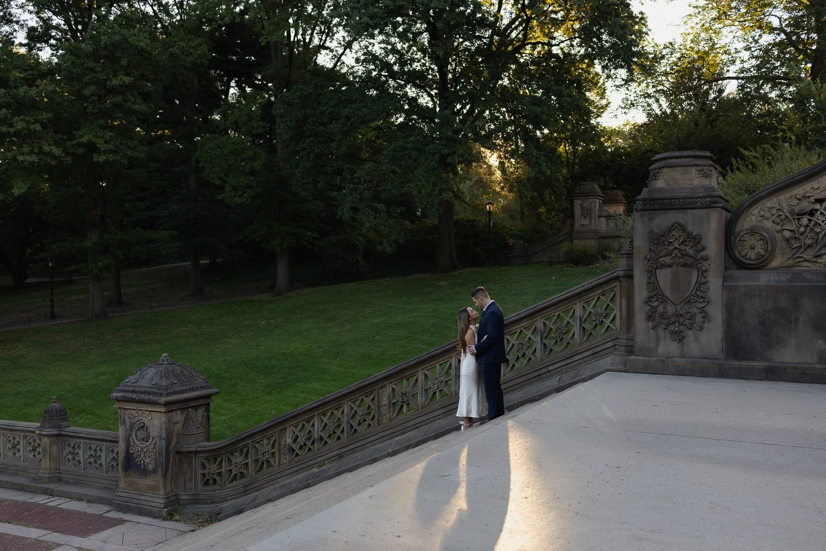 A couple in formal attire standing on a staircase railing in a park during sunset, with trees in the background.