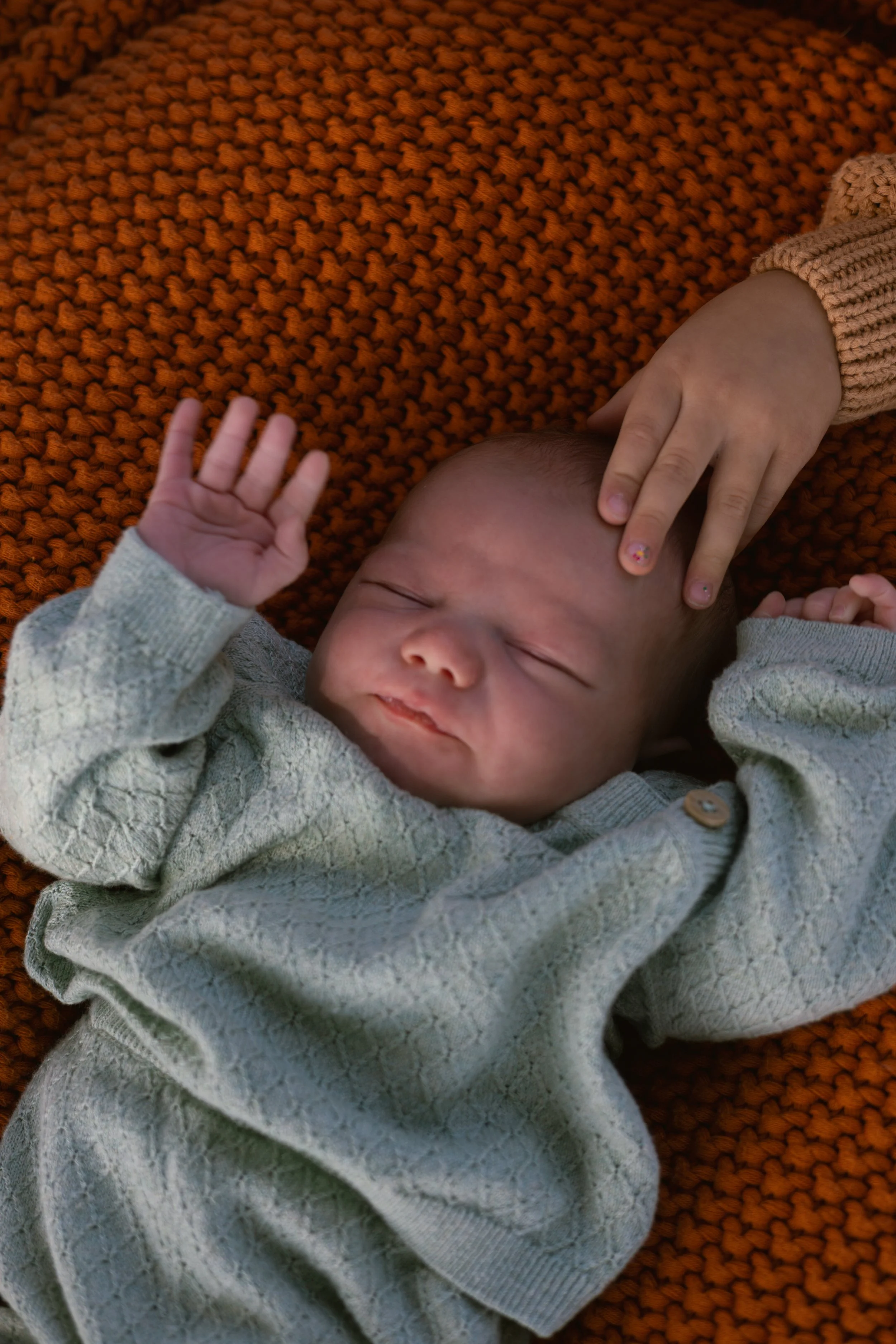 Close-up of a sleeping baby lying on a textured orange blanket, with a child's hand touching the baby's head.