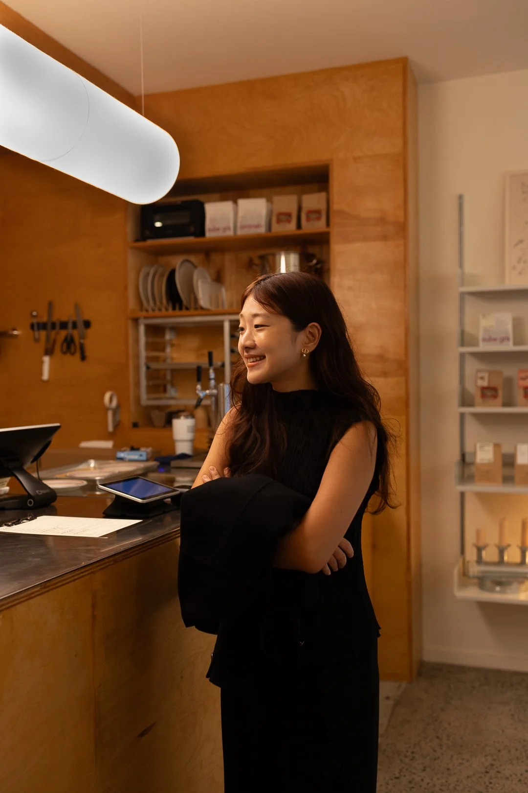 A woman with long dark hair and earrings standing at a counter in a cafe or restaurant, smiling and holding a black coat, with shelves and kitchen utensils in the background.