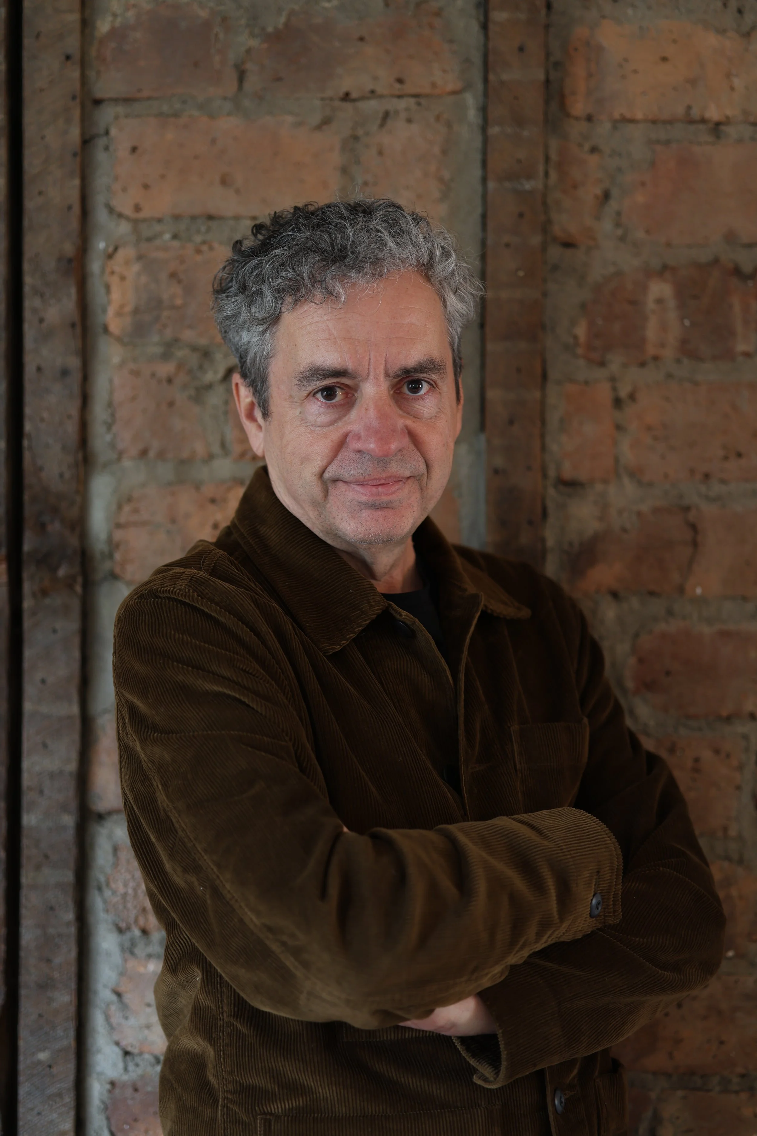 A middle-aged man with gray curly hair, wearing a brown corduroy jacket, standing against a brick wall, arms crossed, looking confidently at the camera.