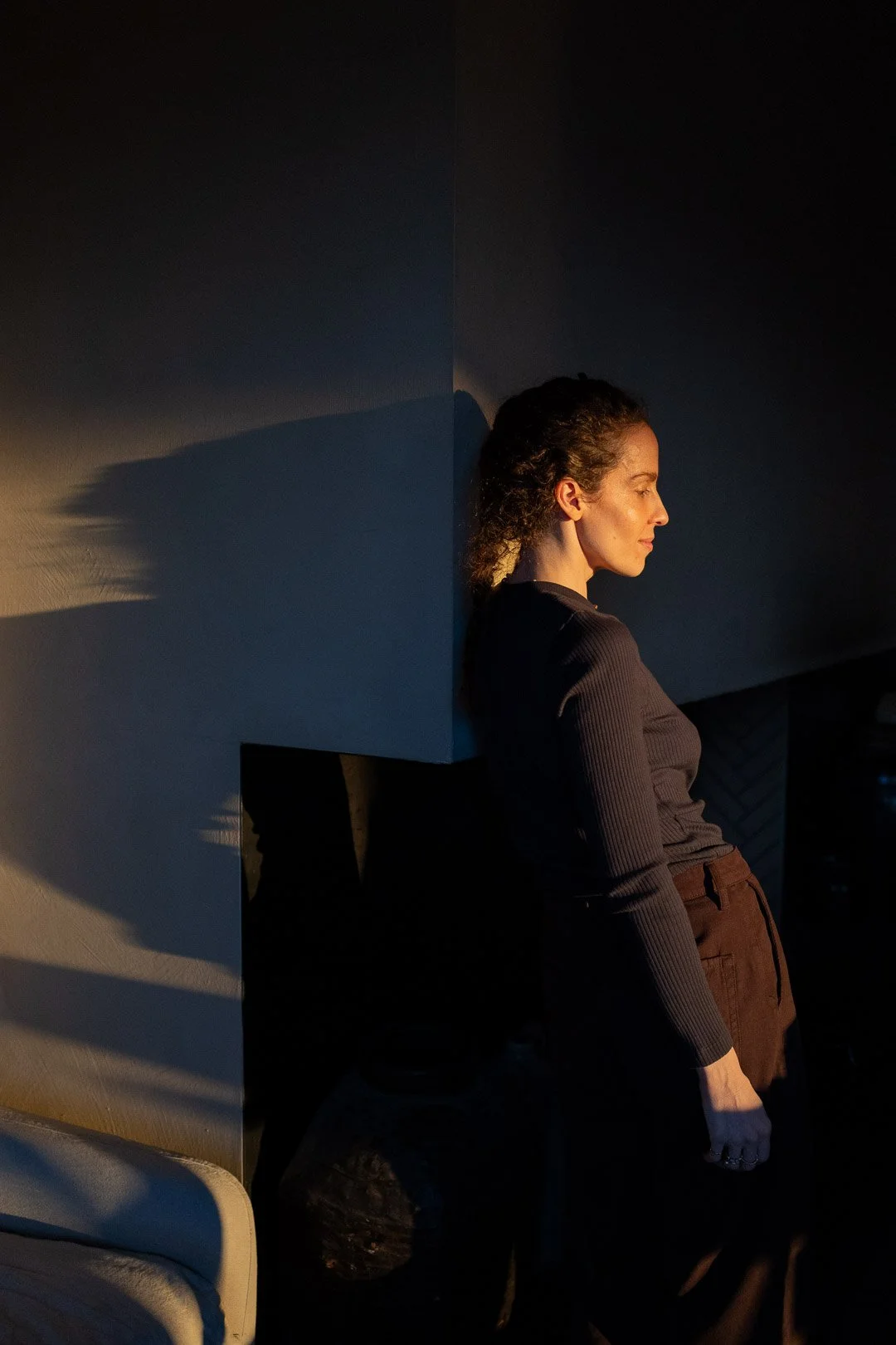 A woman with curly hair standing with her eyes closed, leaning against a dark wall, illuminated by warm sunlight creating shadows.