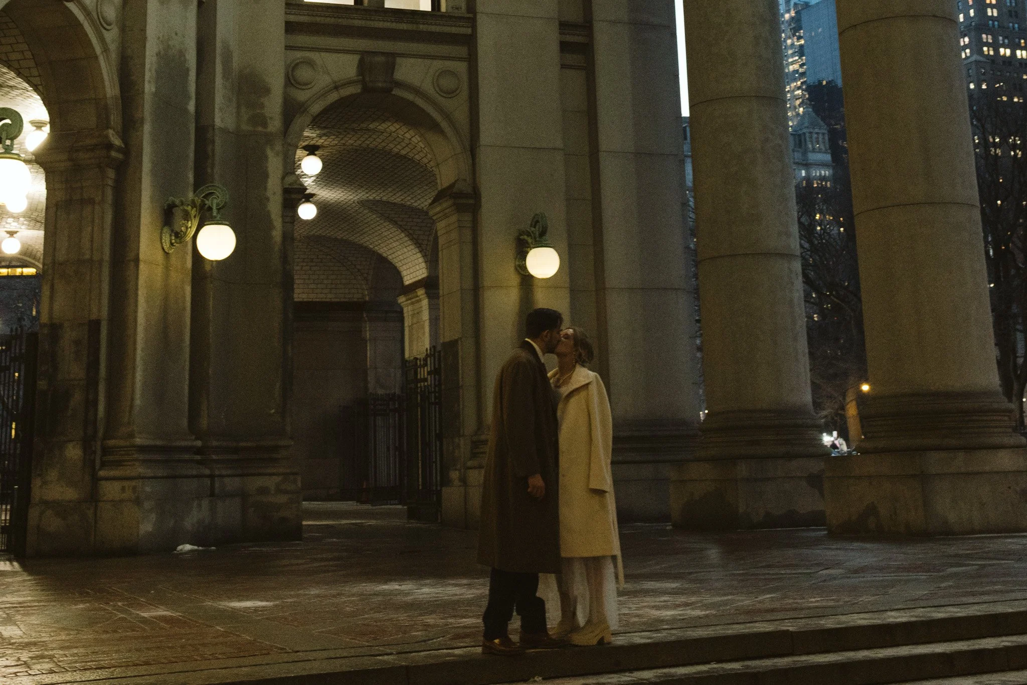 A couple sharing a kiss at dusk outside a historic building with large columns and illuminated spherical lamps.