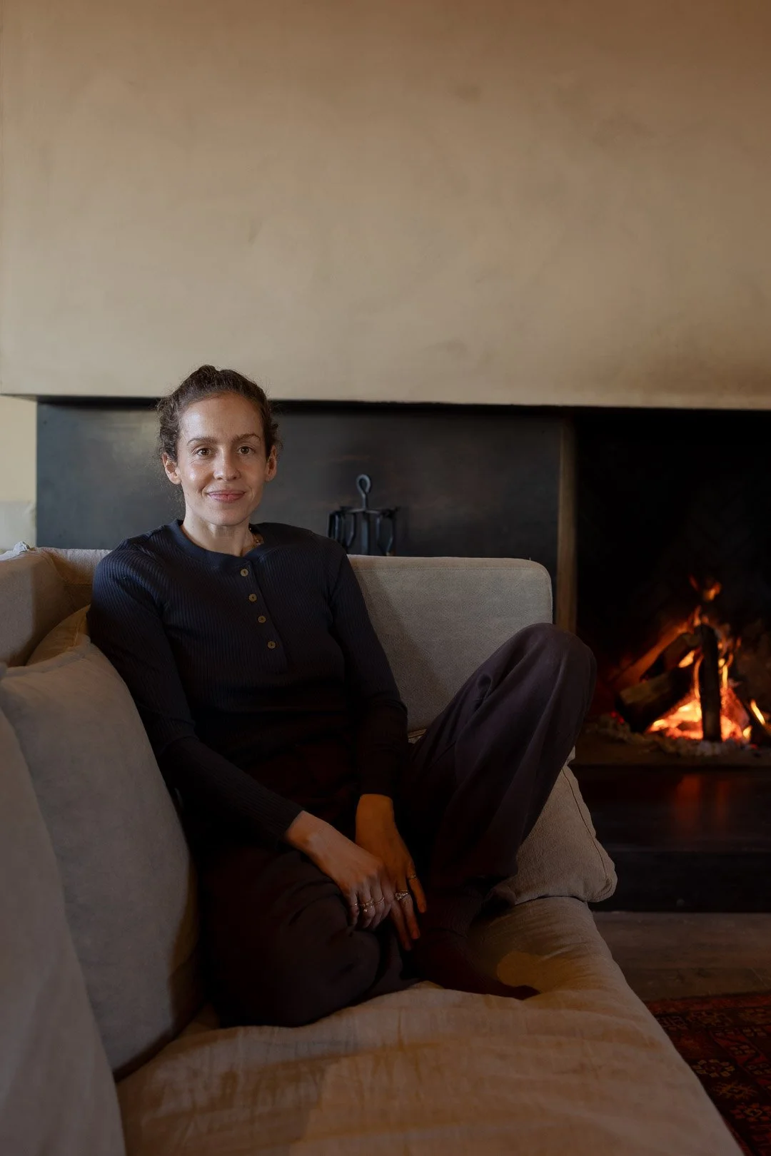 A woman sitting on a beige couch near a fireplace with a fire burning, in a cozy living room.