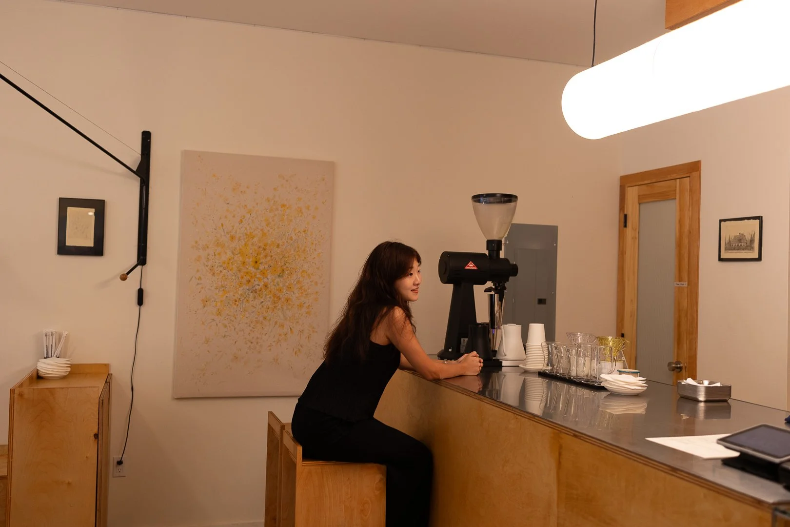 A woman sitting at a coffee bar with a coffee grinder, cups, and glasses, in a minimalist cafe setting with artwork and wooden accents.