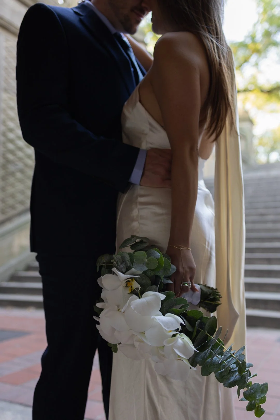 A bride and groom standing close together outdoors, with the groom holding the bride around her waist, and the bride holding a bouquet of white flowers and greenery.