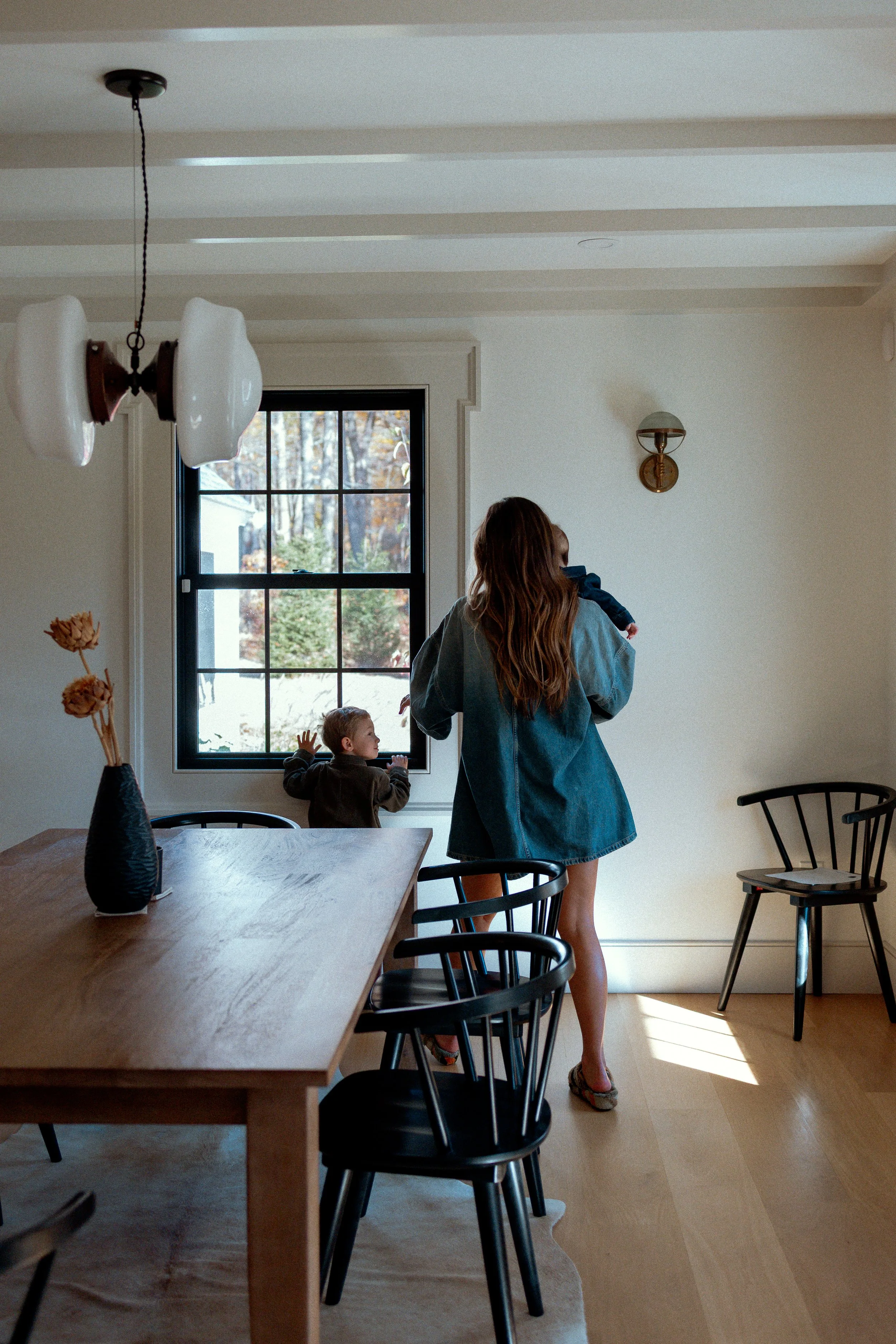 A woman holding a baby walks into a dining room with two children looking out the window. The room has a wooden dining table, black chairs, and a black vase with dried flowers. There is a window with a view of trees outside, and a modern chandelier a