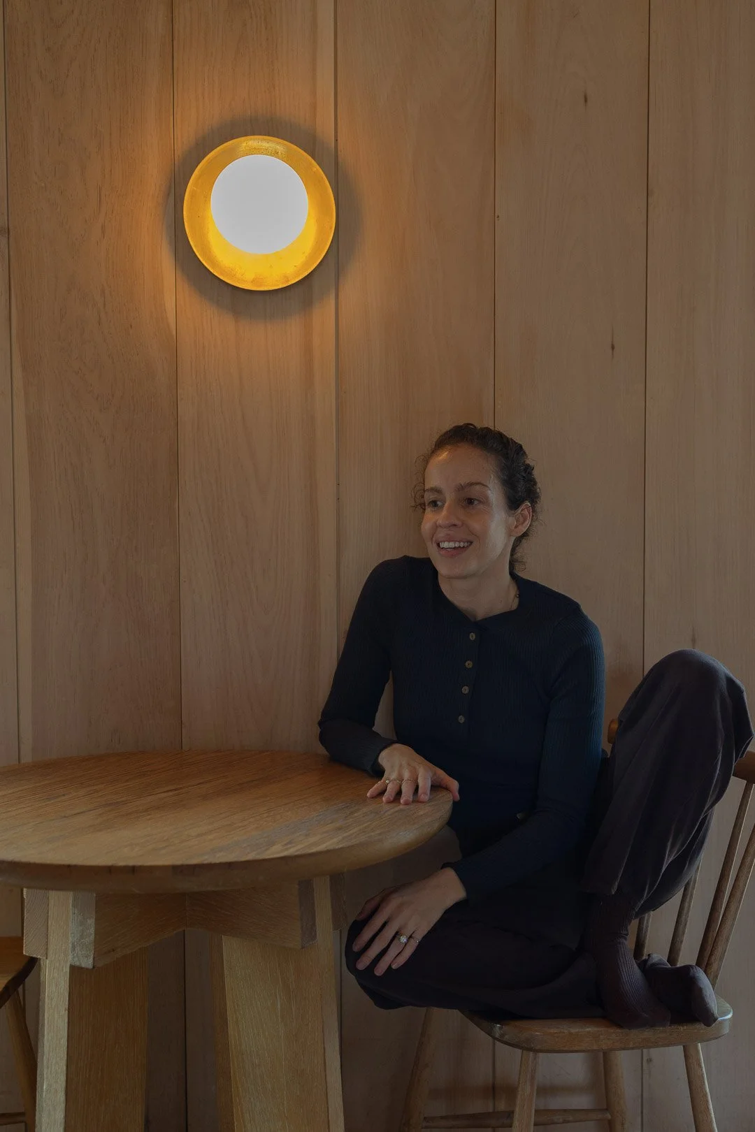 A woman with long, curly hair sitting at a round wooden table in a room with wooden walls. She is wearing a black long-sleeve top and black pants, with a dark coat draped over the back of her chair. There is a yellow circular wall lamp above her on t
