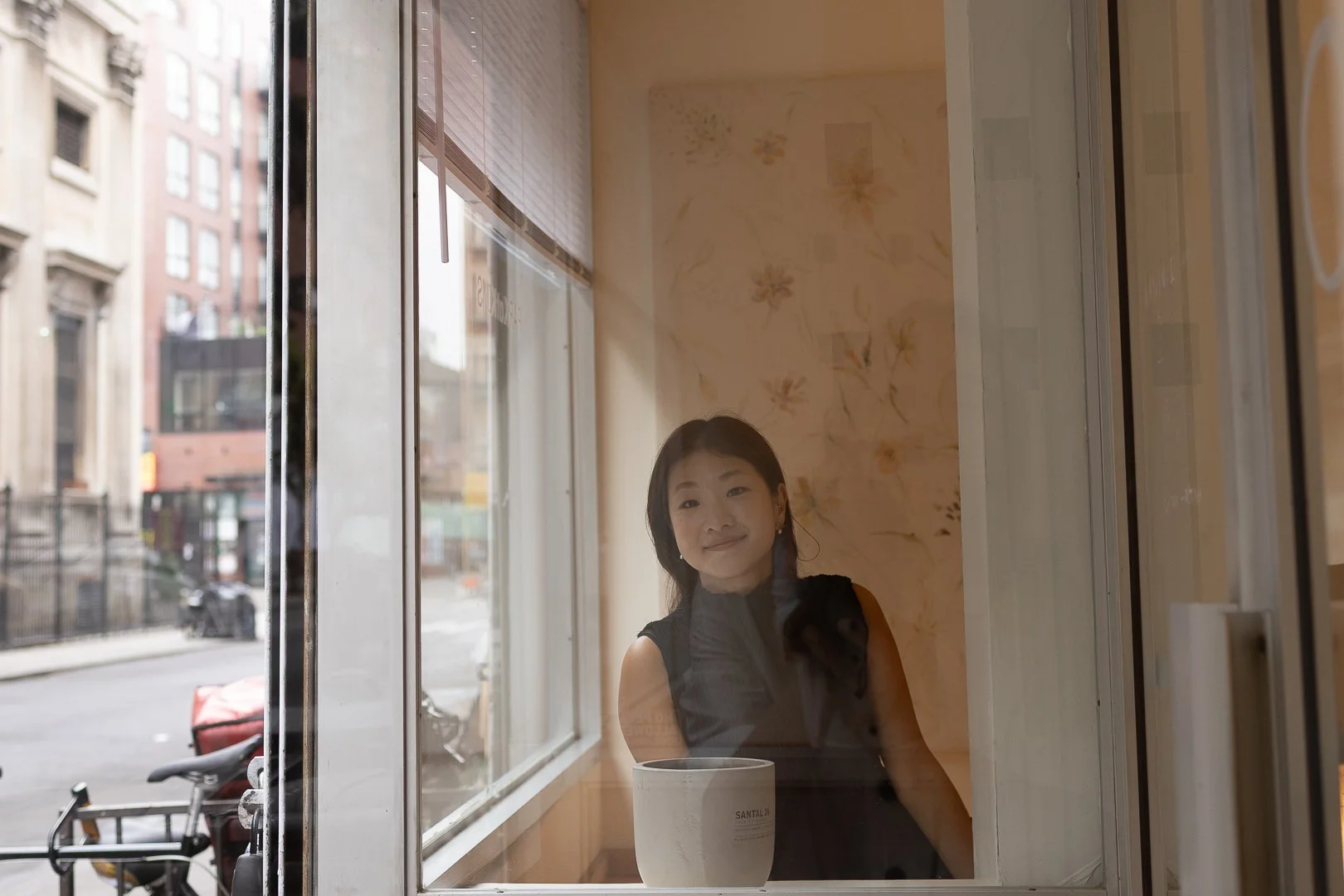 A woman with short dark hair looking out of a café window with a mug on the table in front of her, with buildings and parked bikes outside.