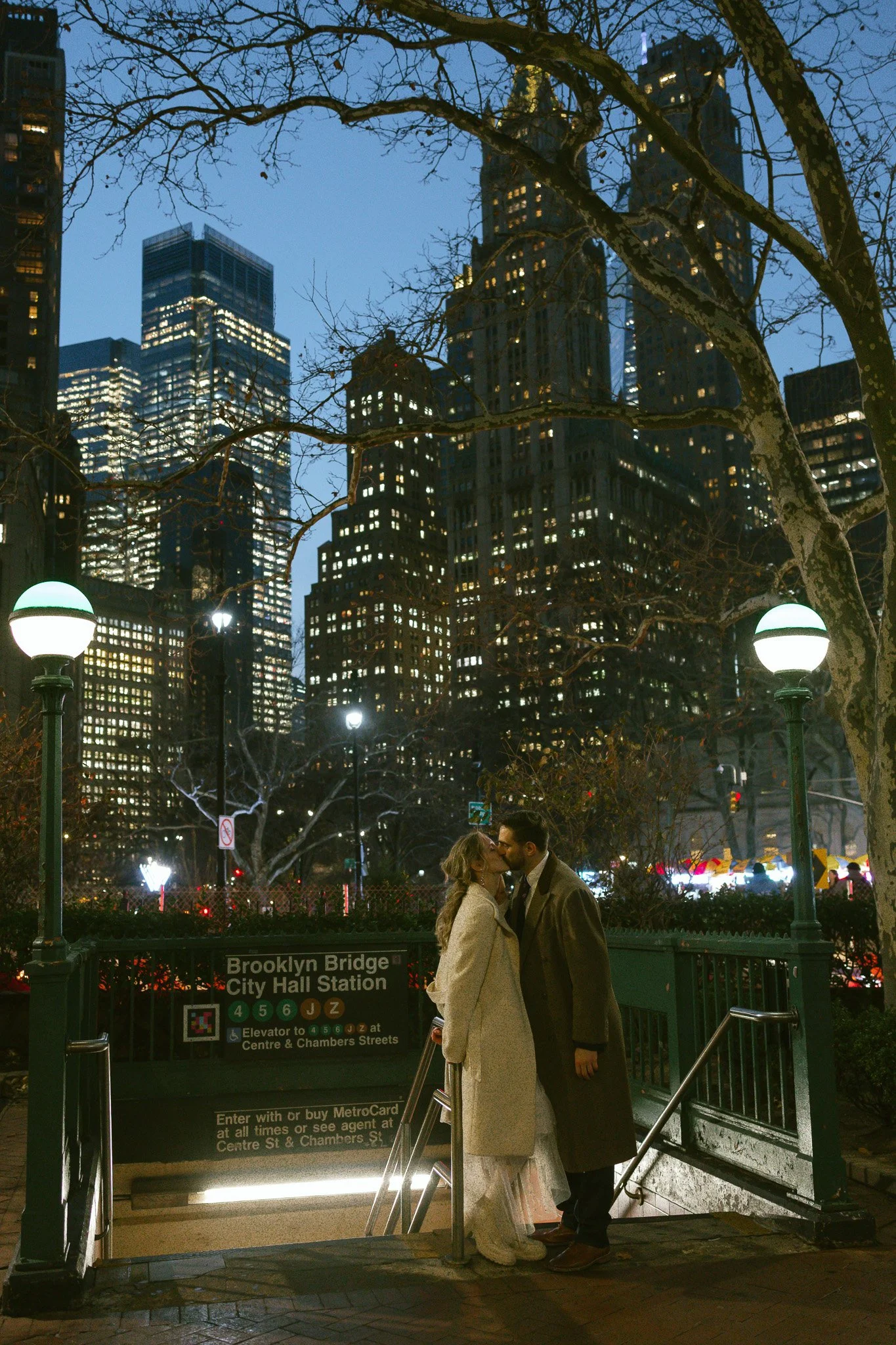 A couple stands kissing at the entrance of Brooklyn Bridge City Hall subway station at night, with tall city buildings illuminated in the background and leafless trees around.
