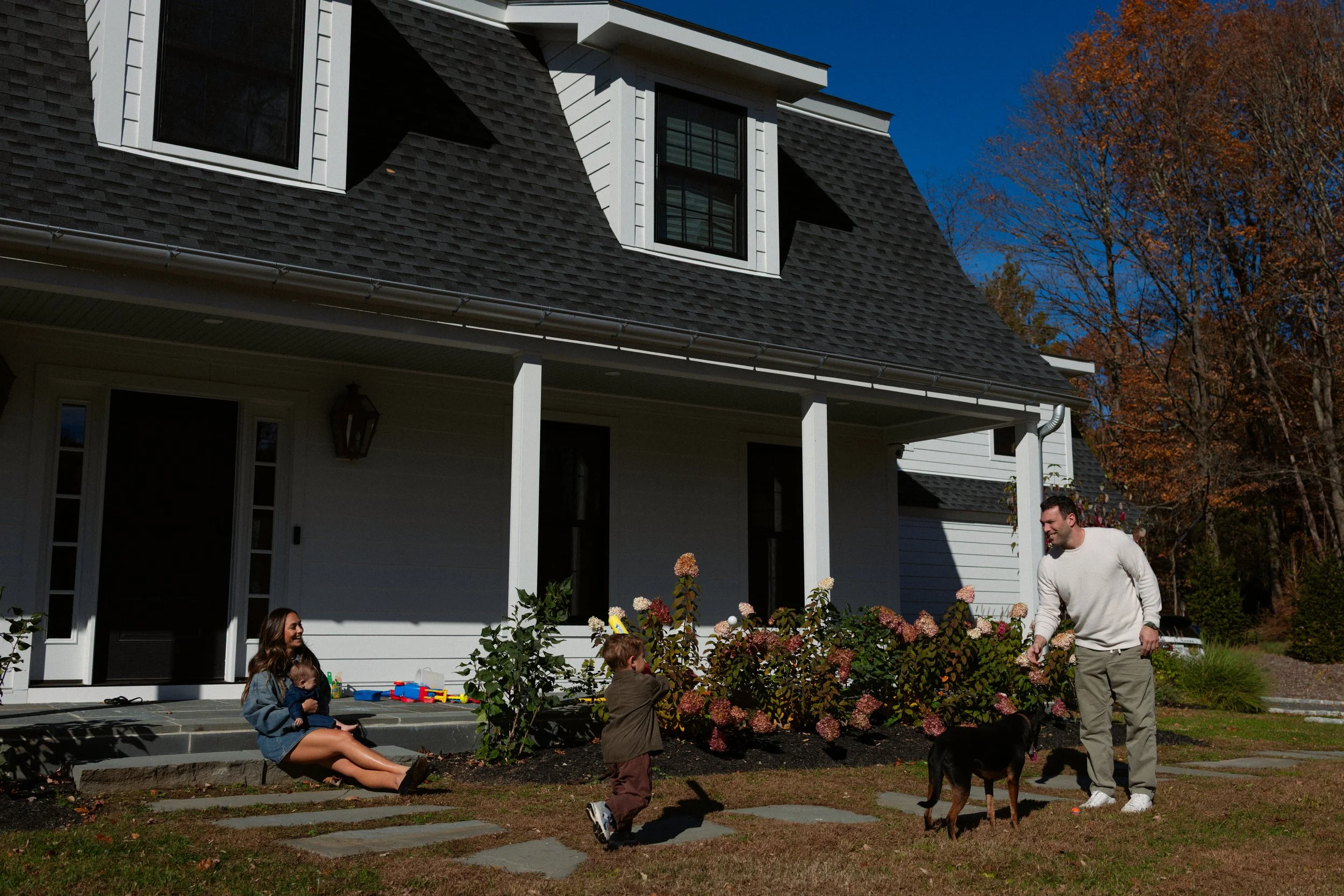 A family of four spending time outside their house on a sunny day. The mother is sitting on the steps with a baby, while a young boy plays with the family dog and the father interacts with the dog. There are colorful toys on the porch and flowering b