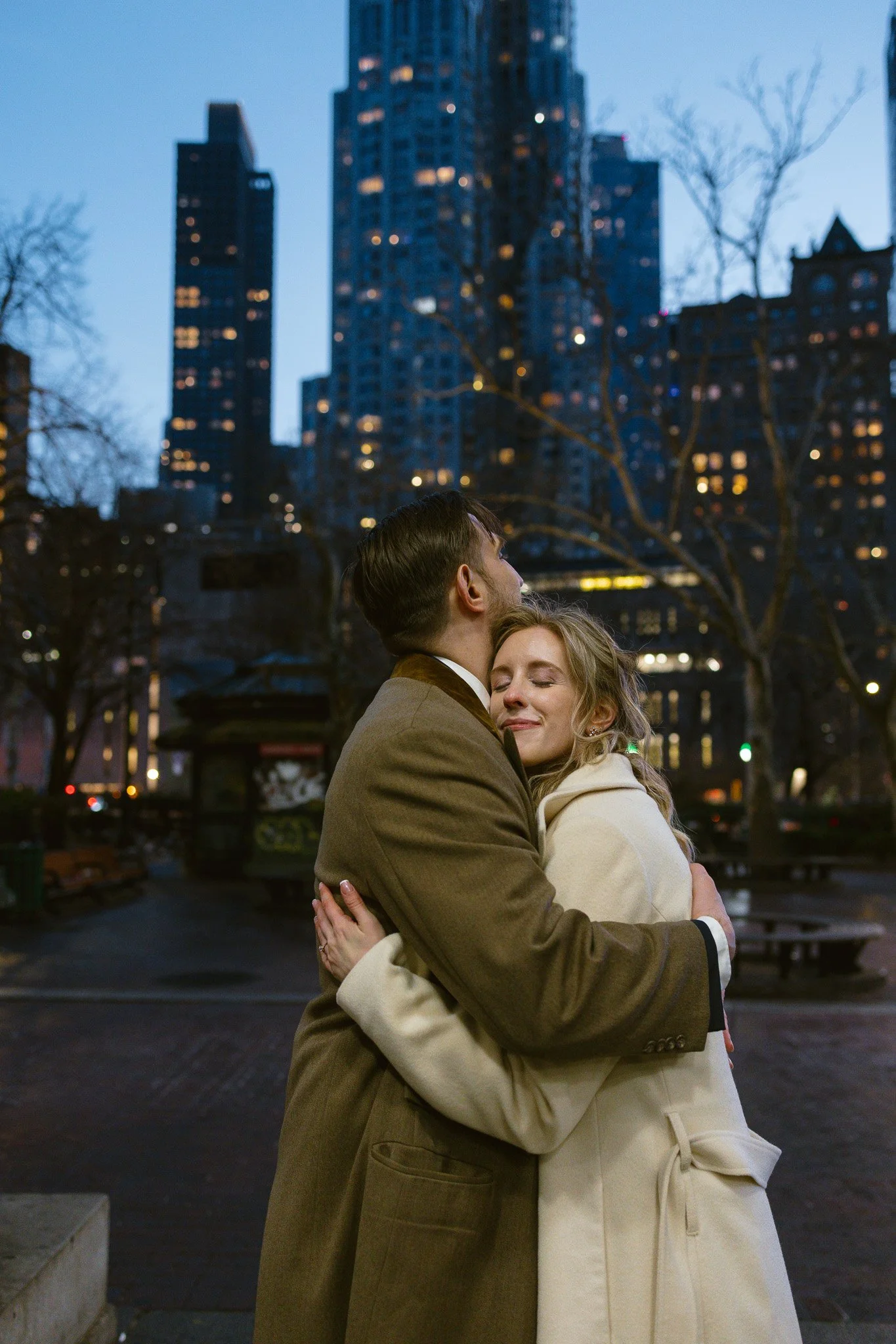 A couple hugging in a city park at dusk, with tall buildings illuminated in the background.