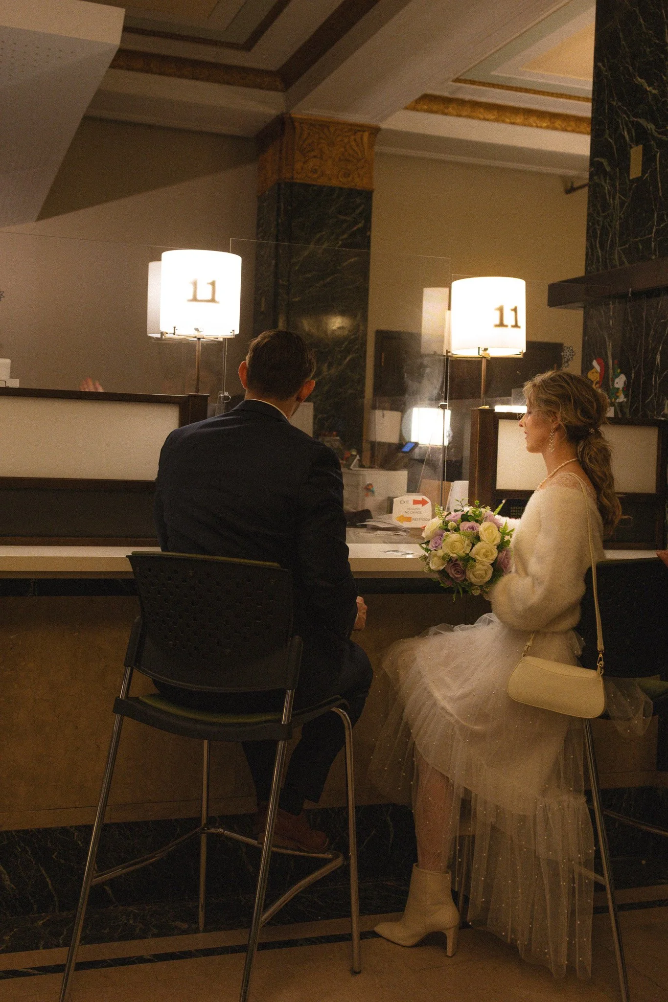 A couple sitting at a hotel reception desk, with the woman holding a bouquet of flowers and dressed in a white dress and a cream sweater, while the man is in a suit, inside a warmly lit hotel lobby.