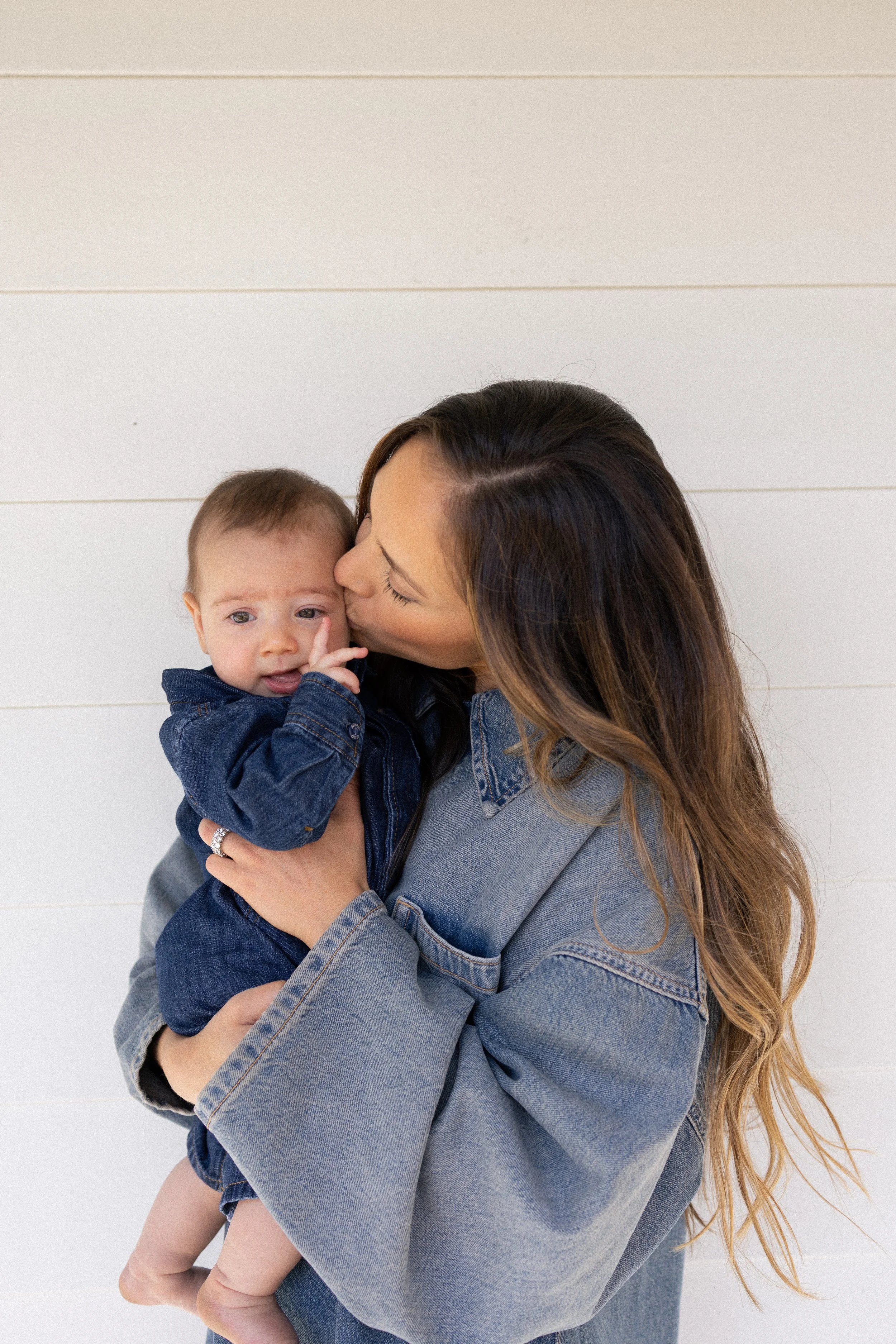 A woman with long brown hair holding a young child in front of a white wall, giving a kiss on the child's cheek as the child looks at the camera and touches their face.