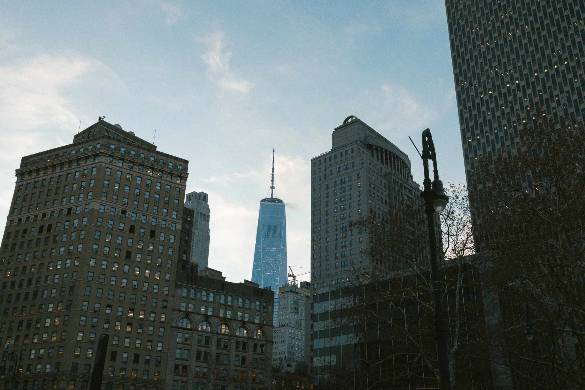 Cityscape of New York City featuring tall buildings, the One World Trade Center in the background, a lamppost, and a cloudy sky.