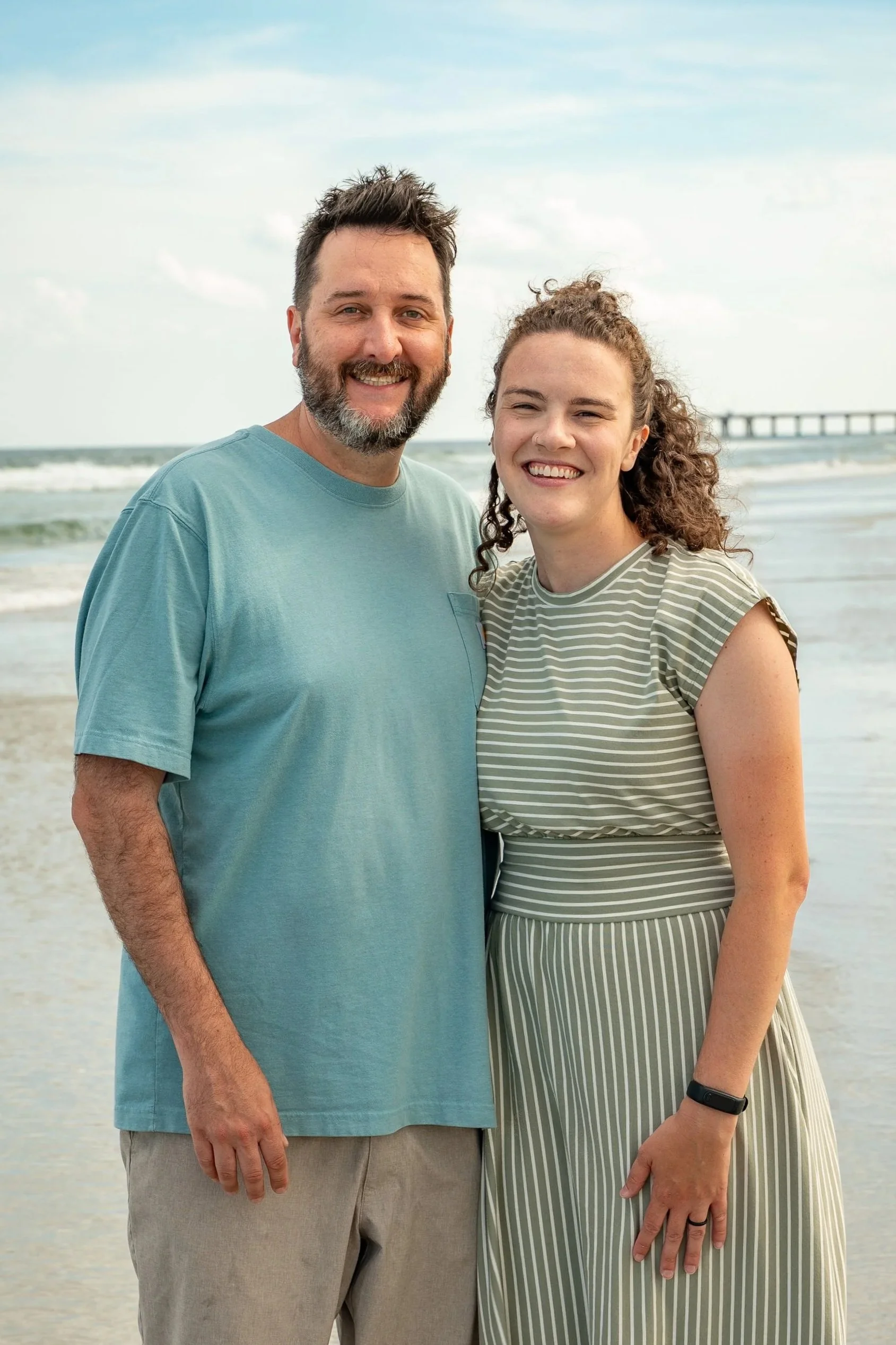 A man and woman standing together on a beach, smiling at the camera. The man has dark brown hair and a beard, wearing a light blue t-shirt, and the woman has curly brown hair, wearing a striped dress. The ocean and a pier are visible in the background.