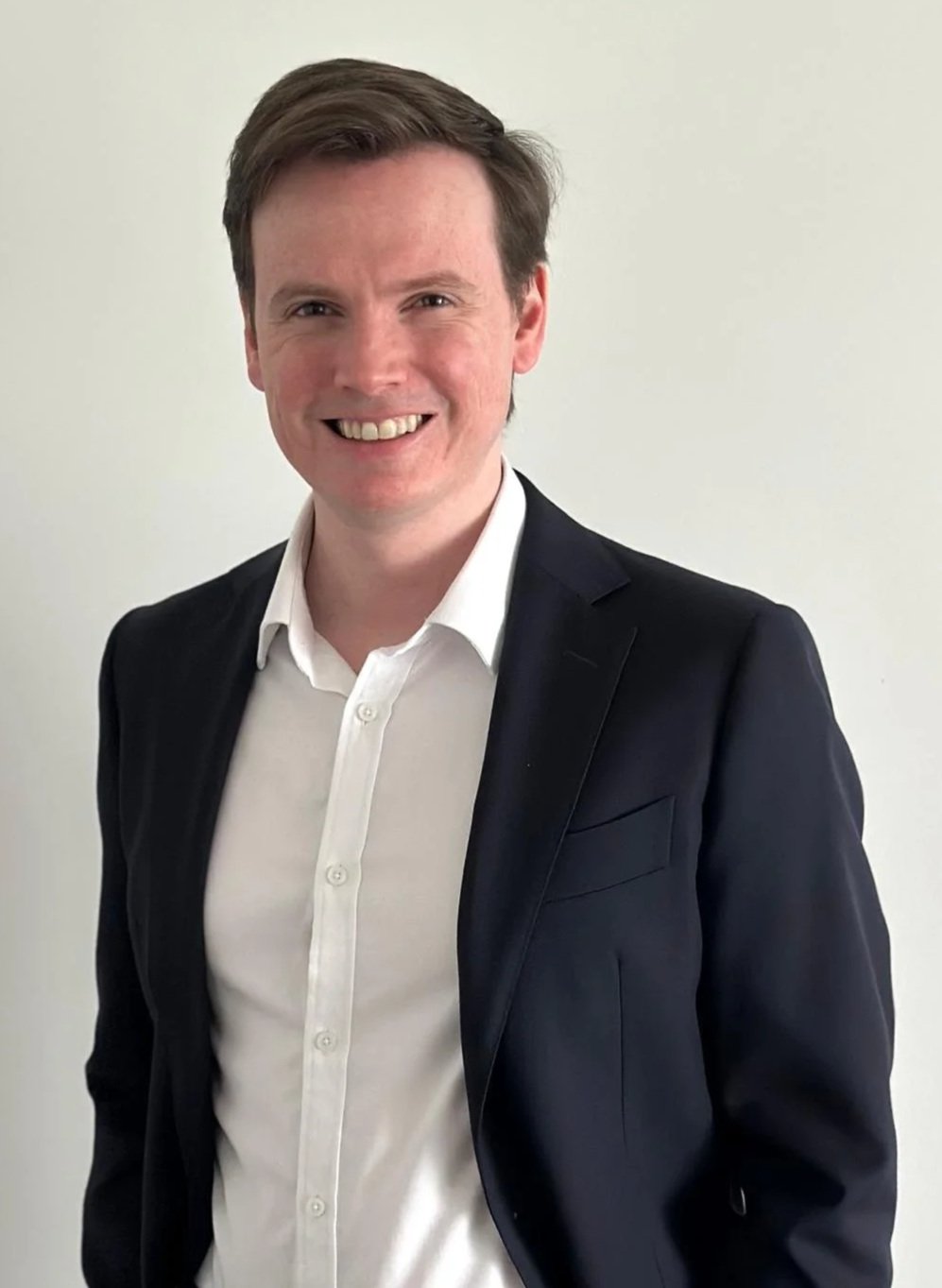 Portrait of a smiling man in a white shirt and black blazer against a plain light background.