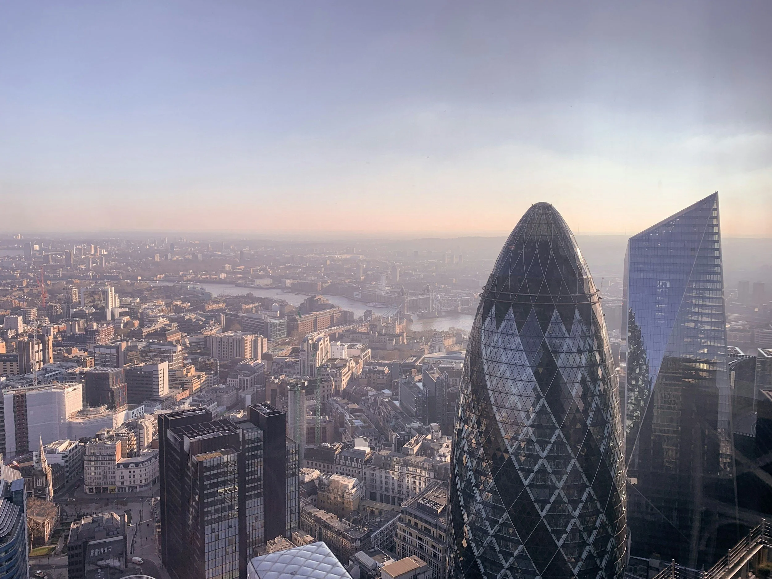 Aerial view of London skyline with prominent skyscrapers, including the Gherkin and tall glass buildings, against a hazy sky. The River Thames is visible in the background.