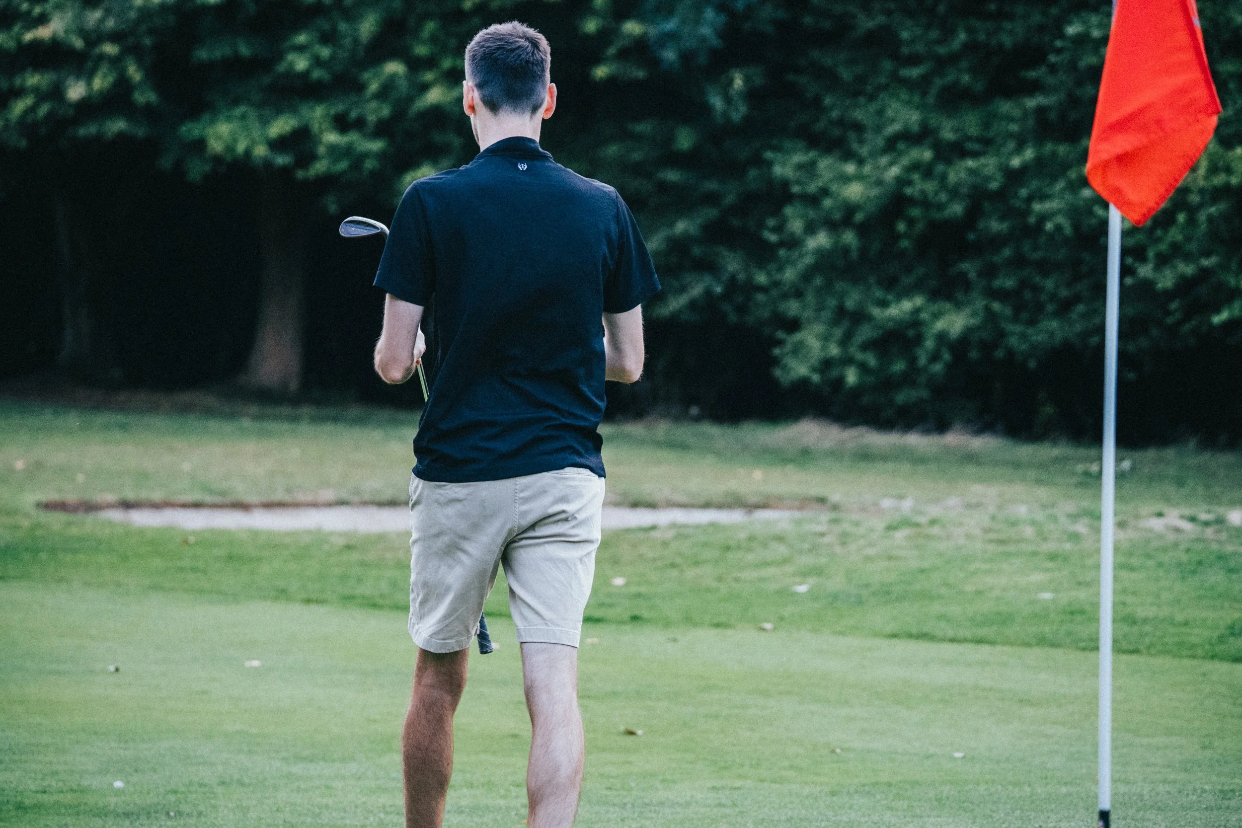 A young man on a golf course preparing to take a shot, with a flag on the green nearby and trees in the background.