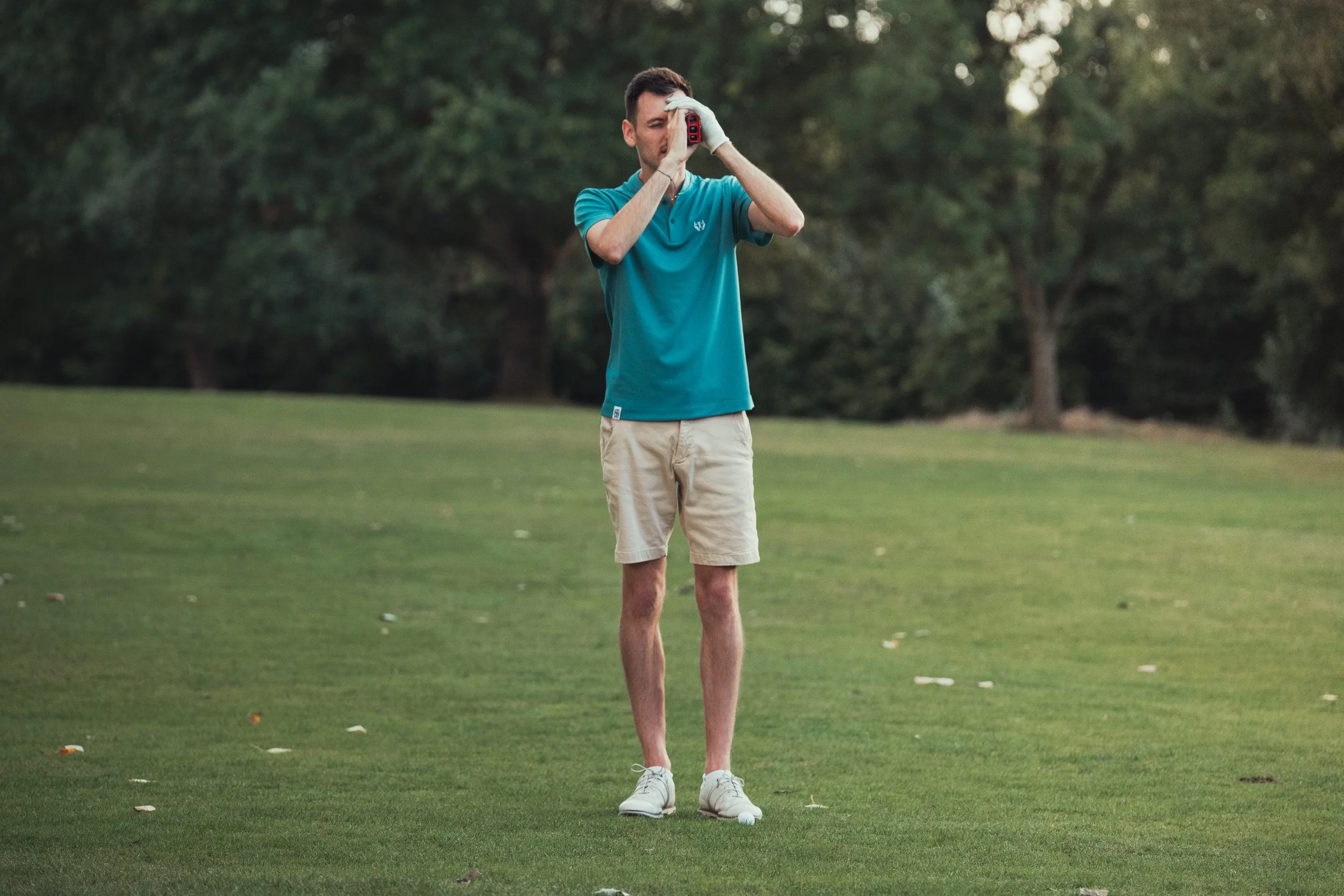 A man standing on a golf course, holding a range finder up to his eye, dressed in a teal polo shirt, beige shorts, and white shoes, with trees in the background.