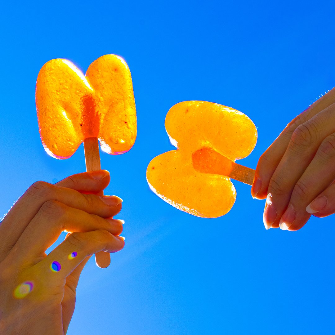 Deux mains tenant des glaces orange aux électrolytes imaginées par le studio d’innovation alimentaire Mâche&Maché, sur fond de ciel bleu.