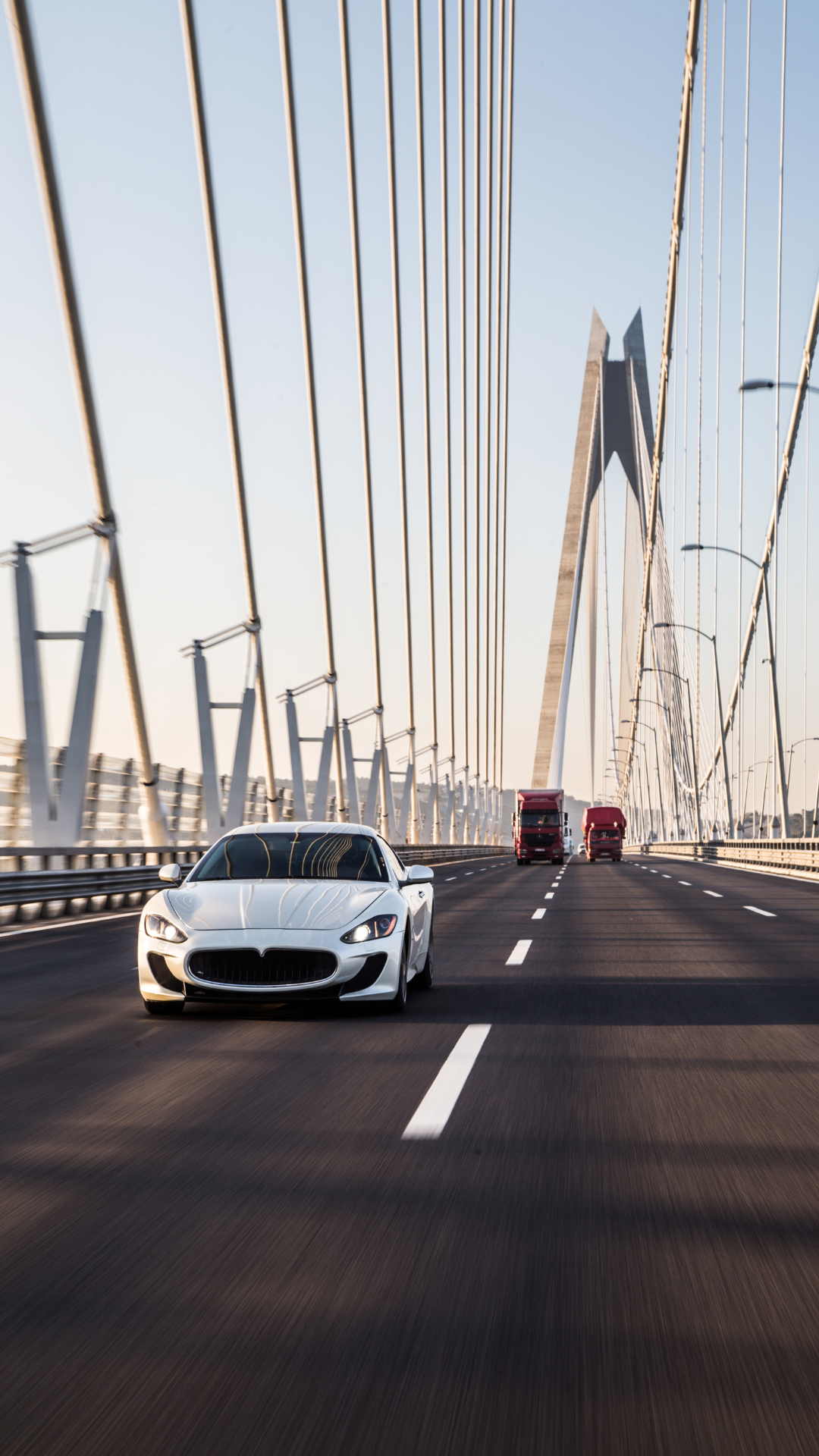 A white luxury sports car with a sleek design driving on a modern cable-stayed bridge, with two red trucks in the background and a clear sky.