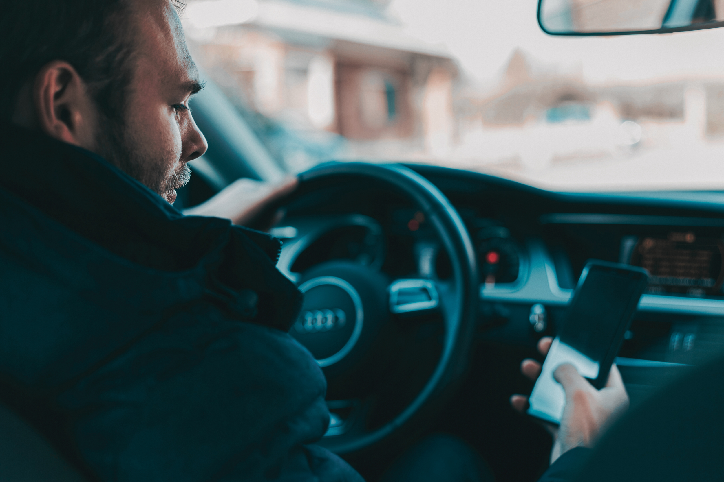 A man with a beard driving a car and looking at his phone, with buildings visible through the car window.