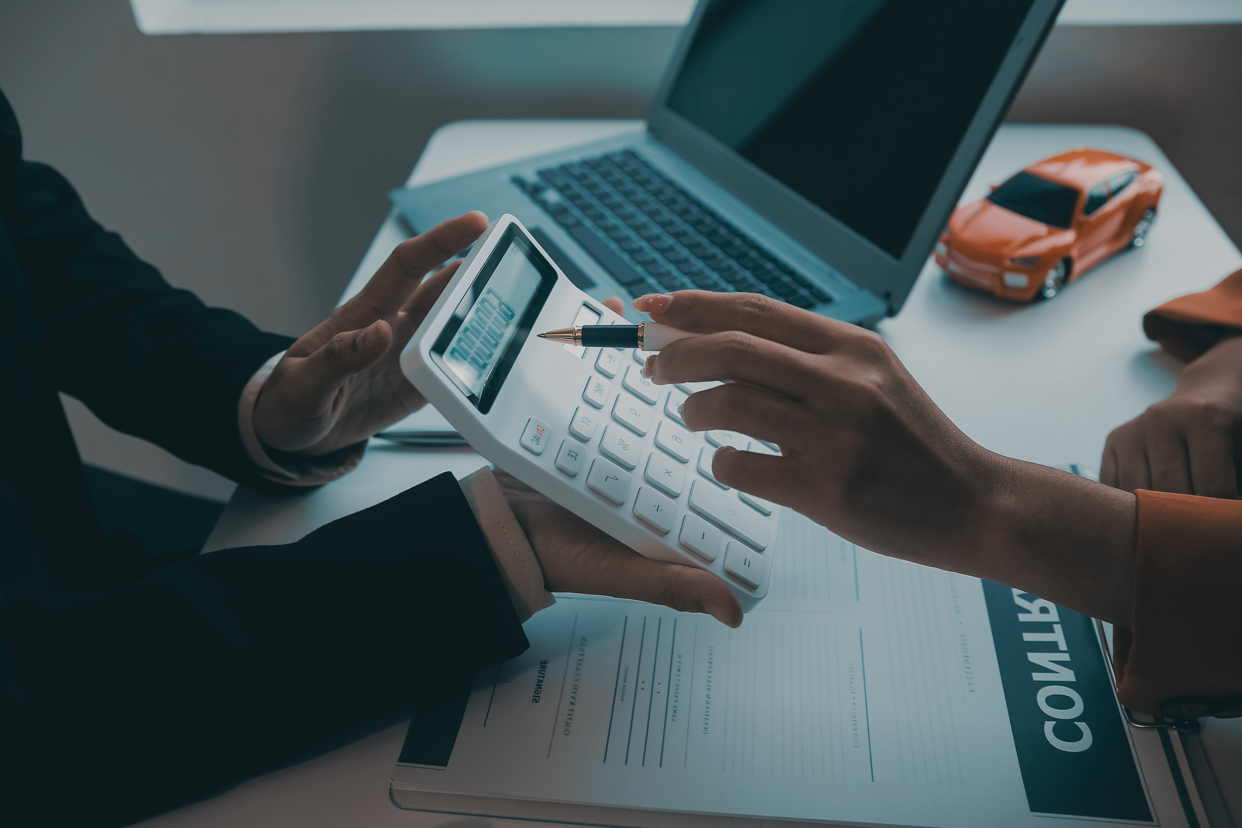 Person holding calculator with a pen, siting at a table with a laptop, a toy car, and documents.