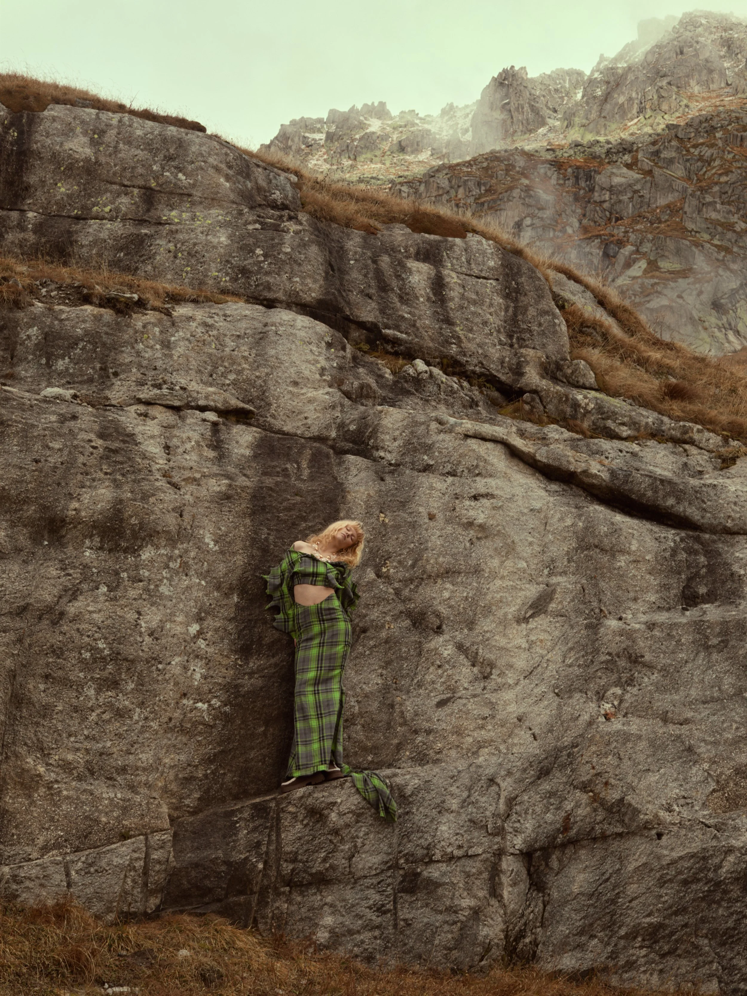 Frau in grüner Klamotte steht auf einer großen Felsformation in den Bergen, hinter ihr nebelige Berglandschaft.