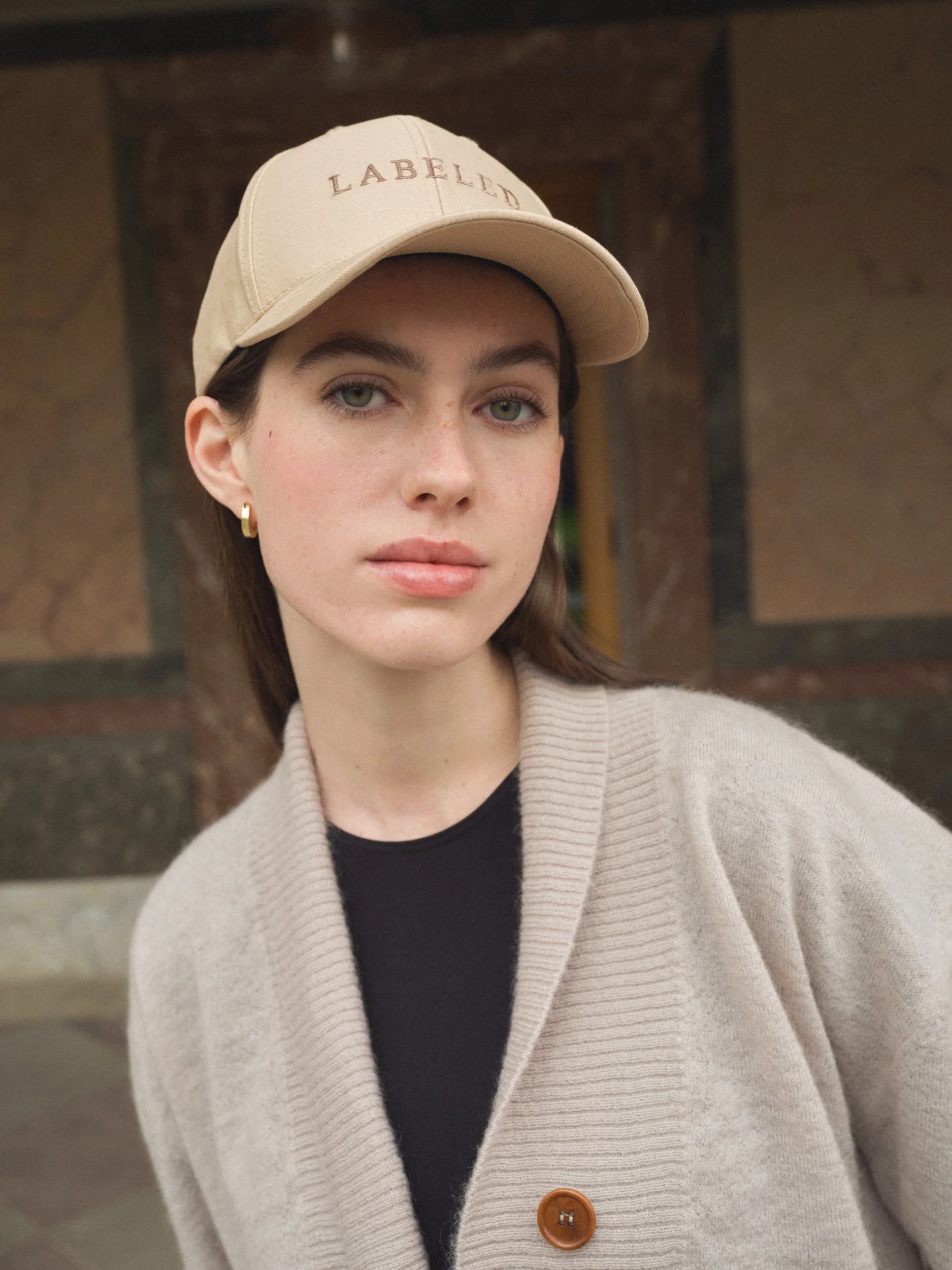 Junge Frau trägt beige Baseballkappe mit aufgesticktem Text und beige Mantel mit braunem Knopf, Hintergrund aus Steinmauer, Blick leicht zur Seite