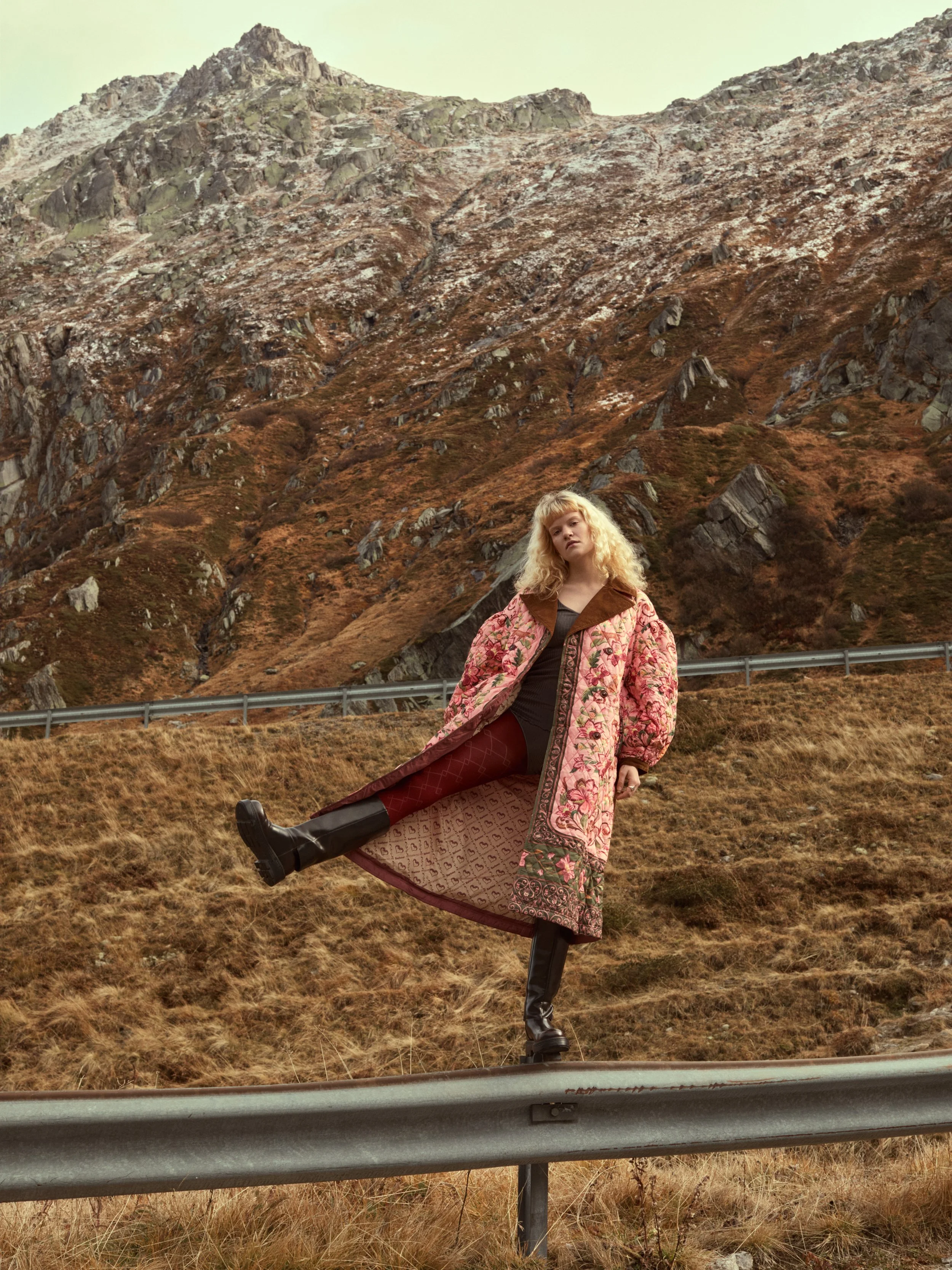 Eine Frau steht auf einem Sicherheitszaun vor einer bergigen Landschaft mit steilen Felsen und trockenem Gras, trägt einen bunten langen Mantel mit Blumenmuster.