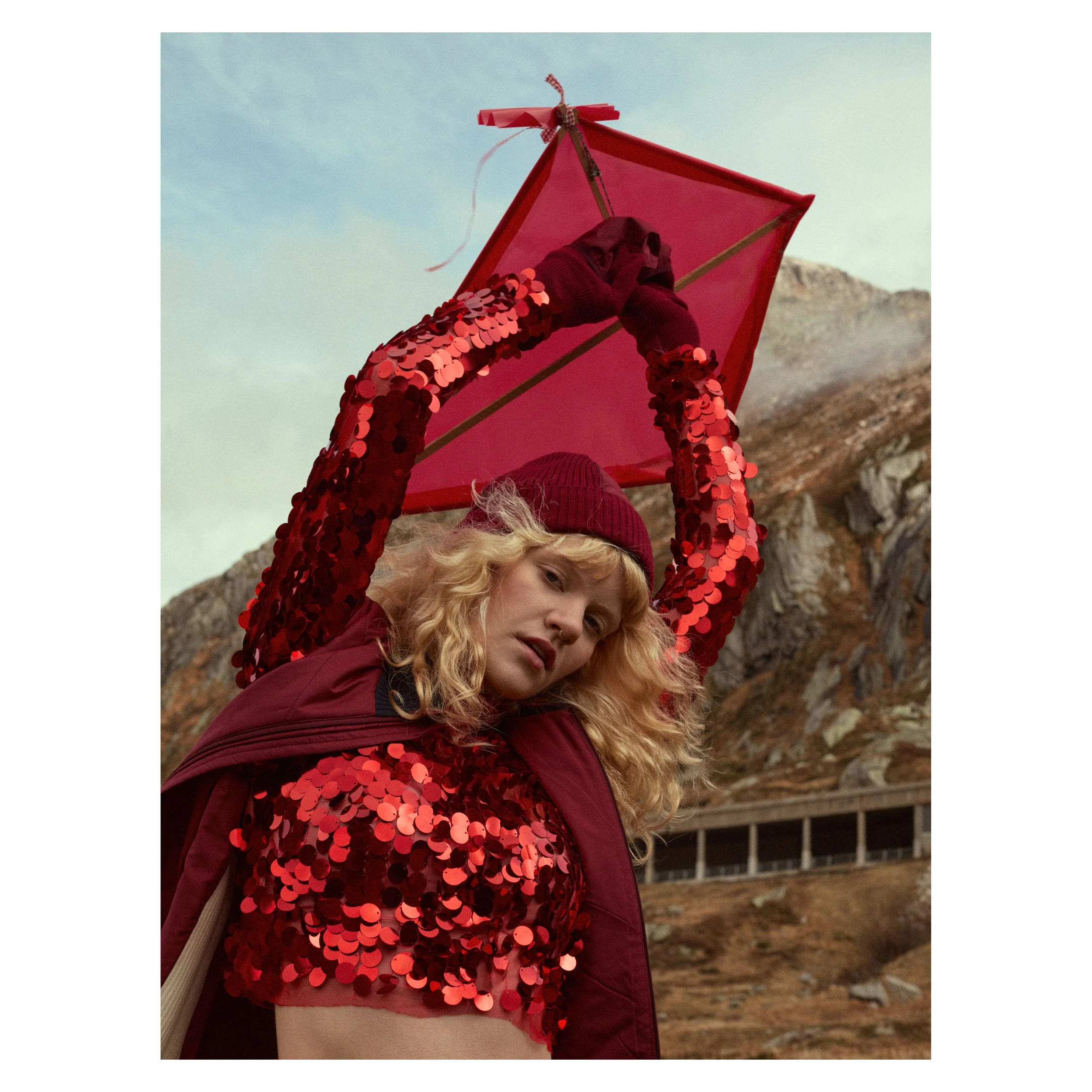 Frau trägt einen roten, Paillettenkleid, einen roten Beanie und einen roten Regenschirm stehen vor einer bergigen Landschaft.