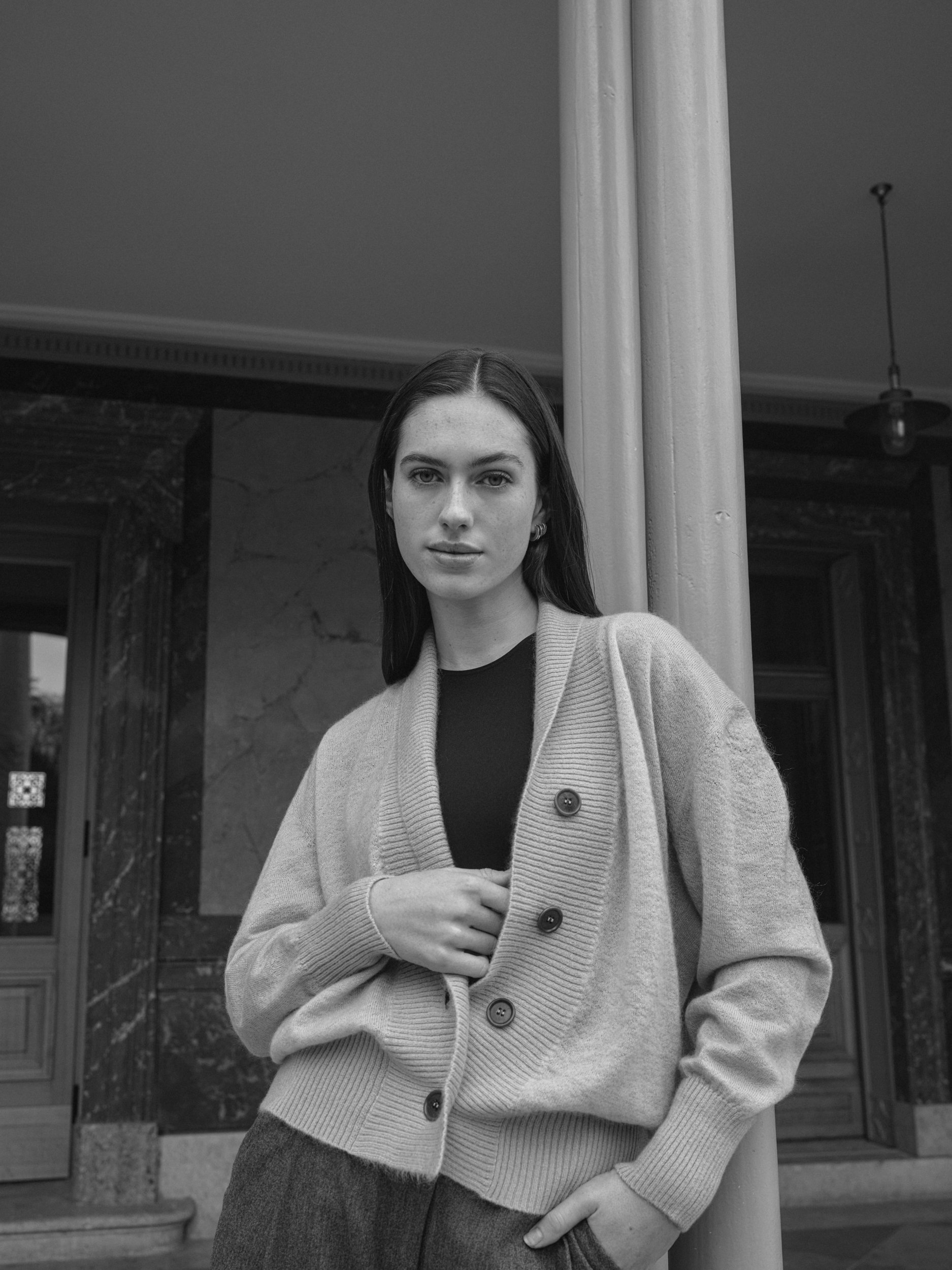 Schwarz-Weiß-Foto einer jungen Frau, die vor einem Gebäude steht. Sie trägt eine Jacke und hat lange dunkle Haare.