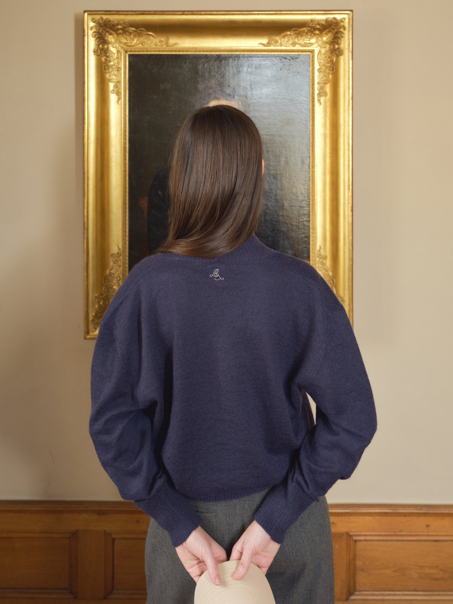 Eine Frau steht mit ihrem Rücken vor einem großen, goldgerahmten Gemälde an einer Wand. Sie trägt einen dunkelblauen Pullover und graue Hosen. In den Händen hält sie einen hellen Hut.