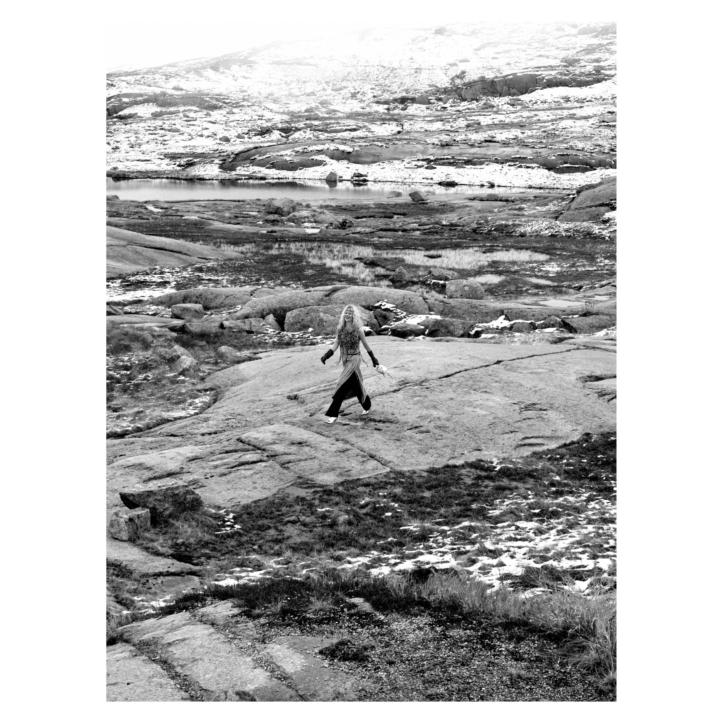 Eine Frau geht auf felsigem Terrain an einem See in einer Gebirgsregion, verschneit und trocken, im Hintergrund Berge und Wasser.