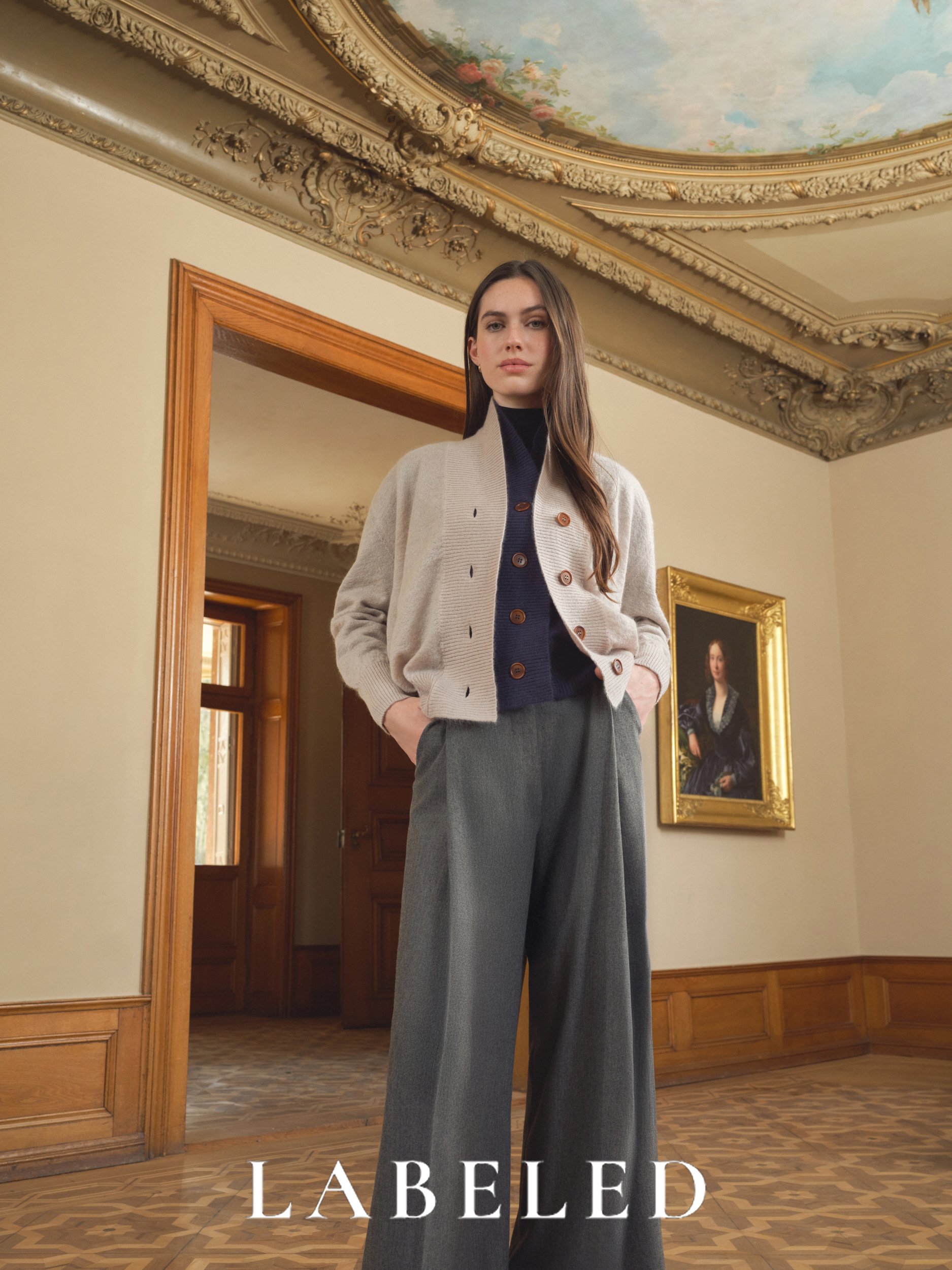 Ein junges Frau in einem eleganten Raum mit kunstvollen Deckenverzierungen und einem porträt im goldenen Rahmen. Sie trägt eine beige Jacke und eine dunkelblaue Bluse, steht vor einer offenen Tür.