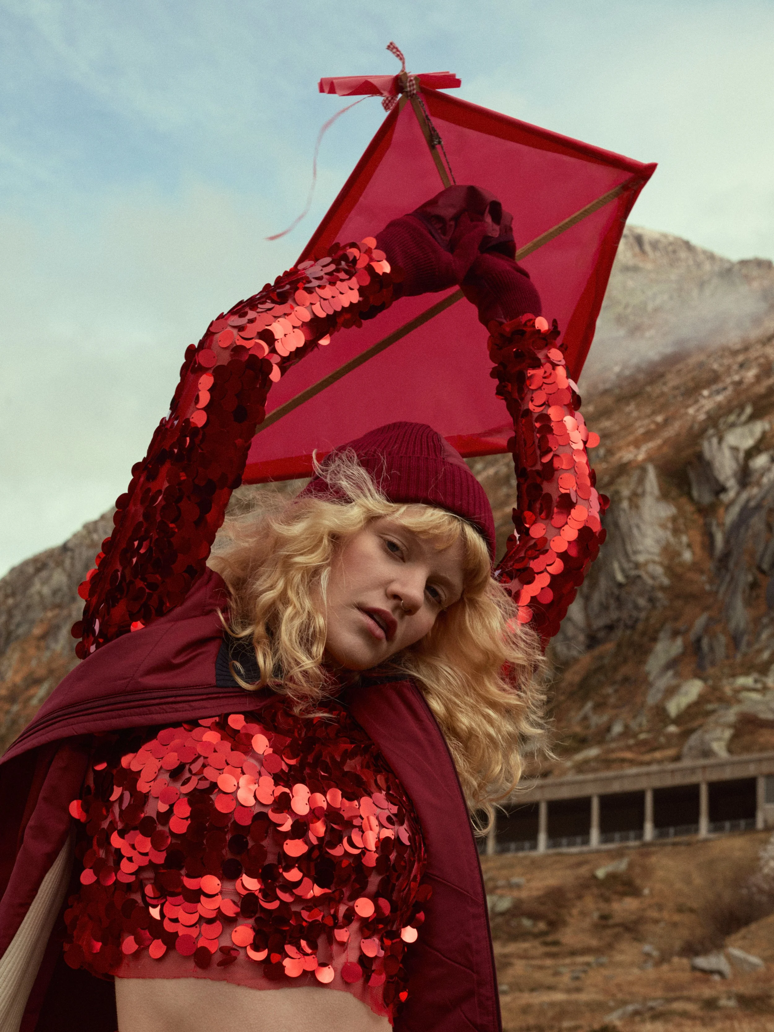 Eine junge Frau mit blonden, lockigen Haaren trägt eine rote Kleidung mit Pailletten und einen roten Hut. Sie hält einen rotem Drachen. Im Hintergrund sind Berge und ein Himmel zu sehen.